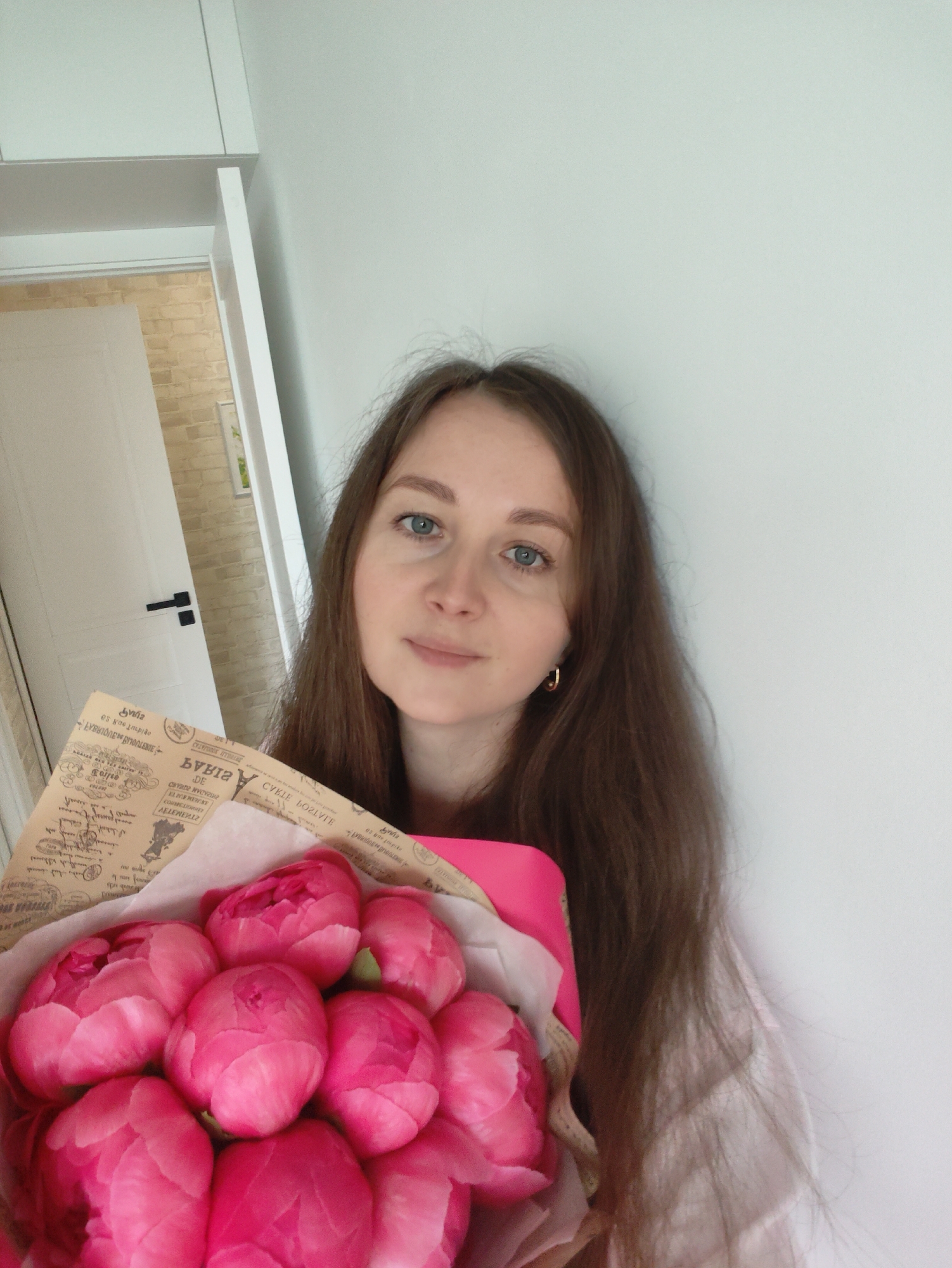 A young woman with long, straight brown hair and light blue eyes poses for a close-up indoor portrait, holding a bouquet of pink peonies. She looks directly at the camera with a calm, pleasant expression. She wears small gold hoop earrings on her right ear.
The vibrant pink peonies, mostly in tight bud form, are wrapped in a rustic brown paper, which features various textual elements and illustrations resembling old newspaper or document clippings. This wrapping paper is layered over a brighter pink fabric or paper.
The background consists primarily of a plain white wall to the woman's right and behind her. To her left, a white interior door with a modern black handle is slightly ajar, revealing a section of a light-colored brick wall and another light-colored surface beyond. Overhead, a white cabinet or shelf structure is visible.
The lighting is soft and even, suggesting indoor ambient light during the daytime.
Visible text on the bouquet's wrapping paper includes: "DE", "Caisse", and several stylized or reversed words such as "CVKLE BOZLVIE" (likely "CYCLE ROSES" or similar, reversed), and "БУКЕТ" (Cyrillic for "Bouquet", reversed). Other text snippets in various languages and fonts, resembling vintage print, are scattered across the paper but are largely illegible.