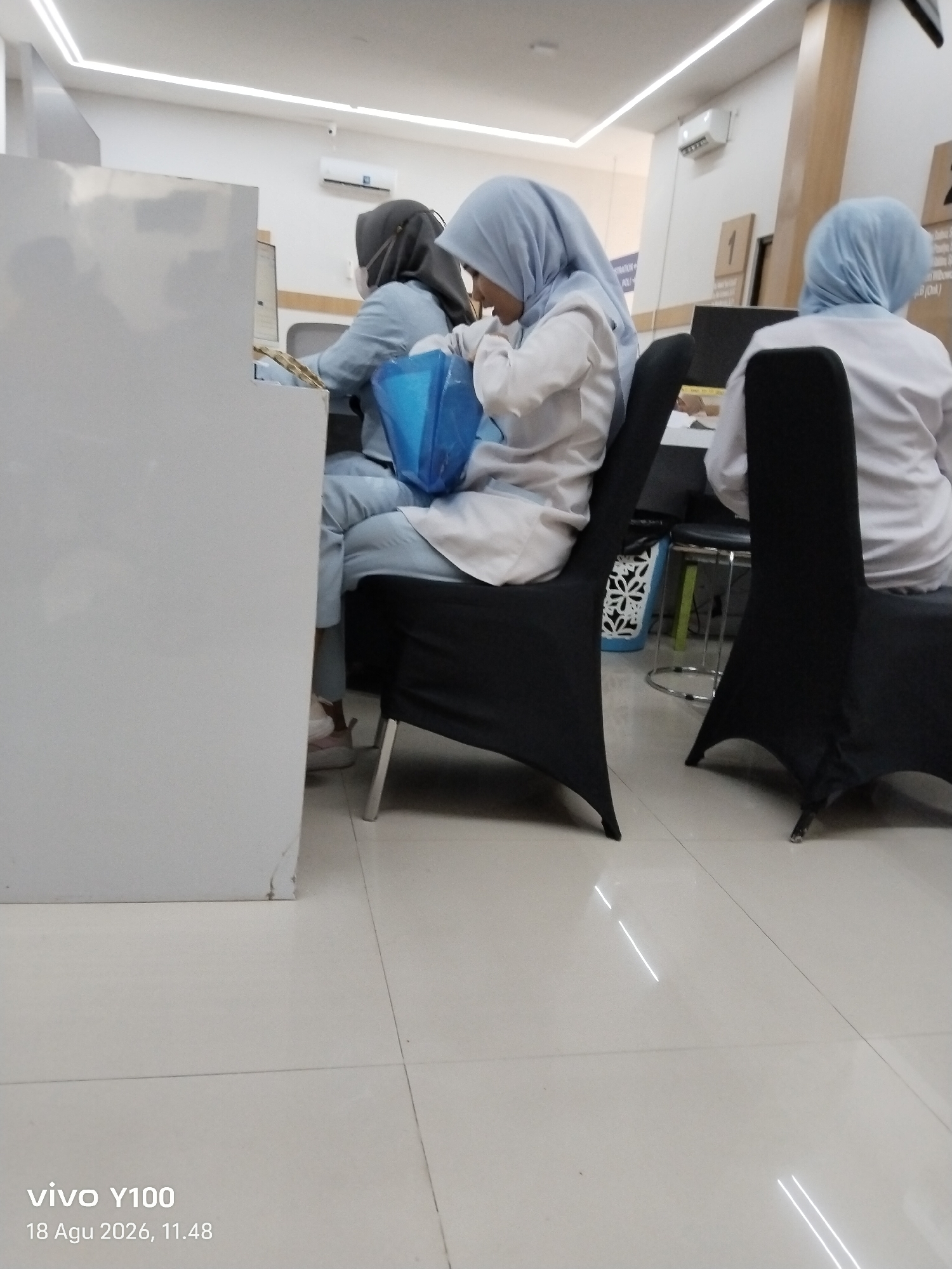 The image shows an indoor scene, likely a waiting area or office, with two individuals wearing light blue uniforms and headscarves sitting at desks. The person in the foreground, on the left, is looking down at a blue plastic bag on their lap, with their right hand inside the bag. They are wearing light pink shoes. The person to their right, further back, is also wearing a light blue uniform and headscarf, and is looking forward, engaged with something on their desk. A third person, partially visible on the right side of the frame, wears a white top and a light blue headscarf and is seated, facing away from the camera. The environment includes white walls, fluorescent lighting, and tiled flooring. A white partition is visible on the left. A sign with the number "1" is visible on the back wall. The "vivo Y100" watermark with the date "18 Agu 2026, 11.48" is present in the bottom left corner. While the image is undated and does not explicitly show the city of Patokan, Indonesia, it depicts a common administrative or healthcare setting that could be found in such a location.