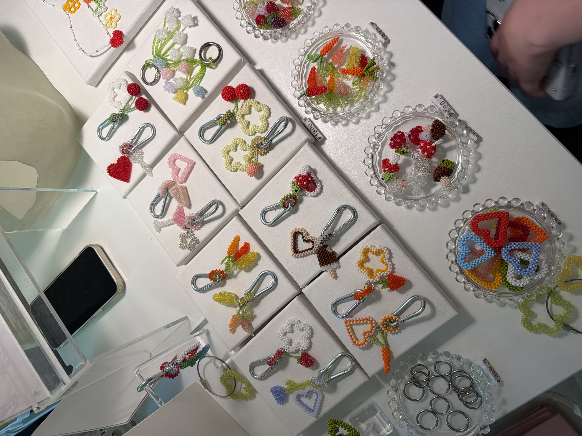 The image shows an assortment of handcrafted beaded keychains on display. The keychains are presented on small, square, white platforms and clear decorative dishes, arranged on a white surface that appears to be a retail display table. The keychains exhibit a variety of shapes and colors, including hearts, flowers, fruits, and other whimsical designs. Each keychain features a silver-toned metal clip. A smartphone is visible in a clear display case on the left side of the image. A person wearing a light blue shirt is partially visible on the upper right side, holding a smartphone and taking a picture. The scene appears to be in a retail store. The stickers with prices written in the Russian language (currency is indicated by the symbol P) suggest that the location is in Moscow, Russia.