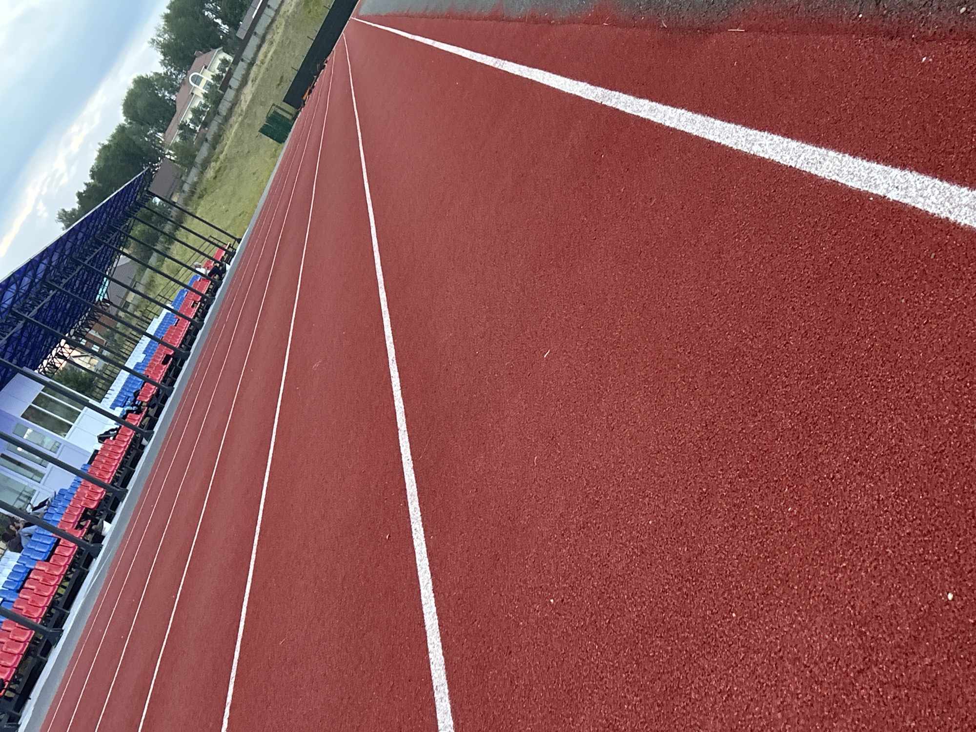 The image shows a red running track at an outdoor stadium. The track has white lane markings. In the background, there are bleachers with red and blue seats, and a building with a blue roof. Behind the stadium, there are houses and trees. The sky is cloudy. This is a scene from the city of Noviy, Russia. There are no visible actions or interactions.