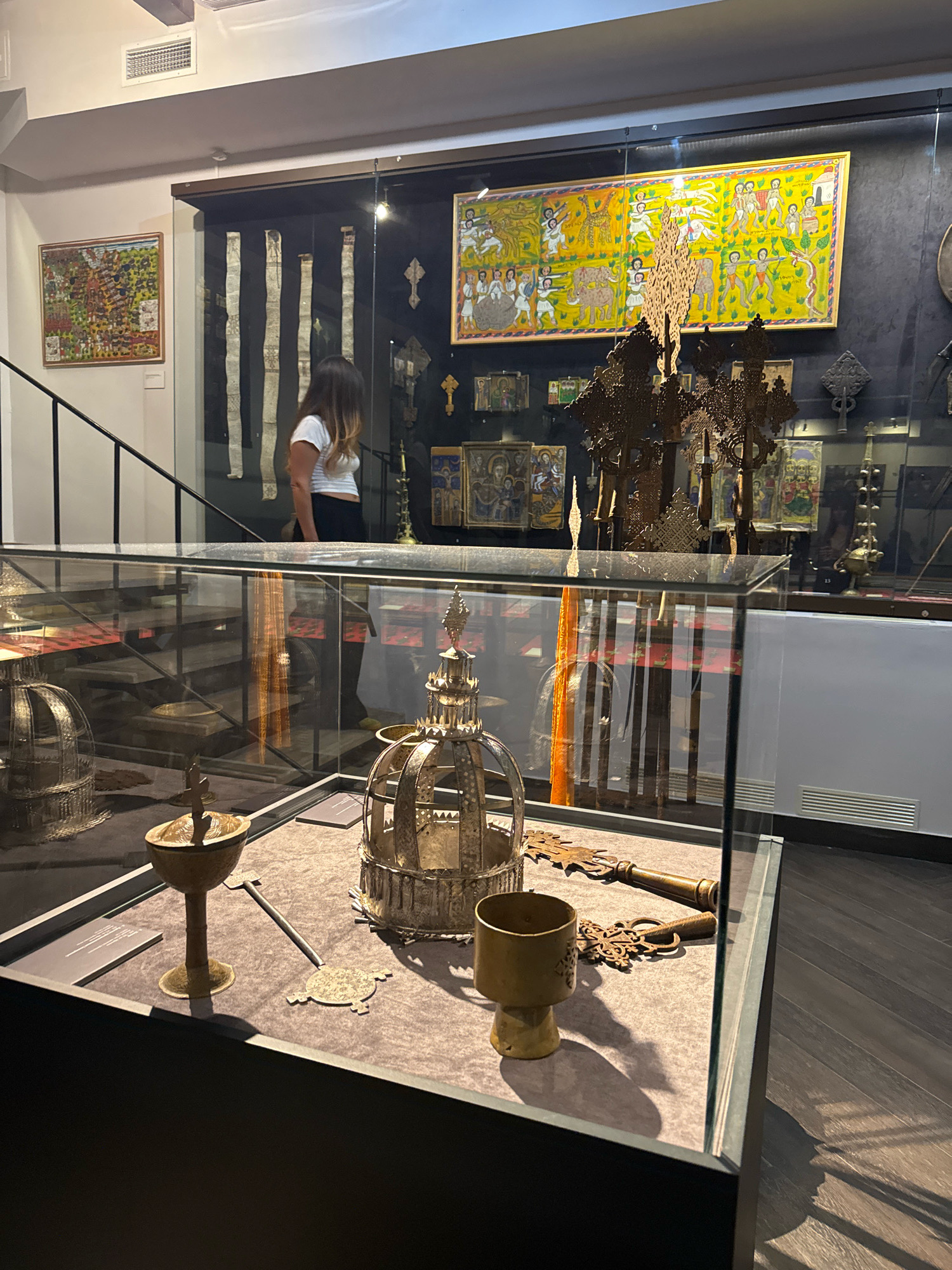 The image shows a display case in a museum. Inside the case are several religious artifacts, including crosses, a chalice, and a key. The artifacts are made of metal and appear to be old. In the background, there are paintings on the wall, as well as a woman looking at the display. The museum appears to be well-lit, with spotlights illuminating the artifacts. The floor is made of dark wood. The overall impression is one of reverence and historical significance. The museum is located in Moscow, Russia.
