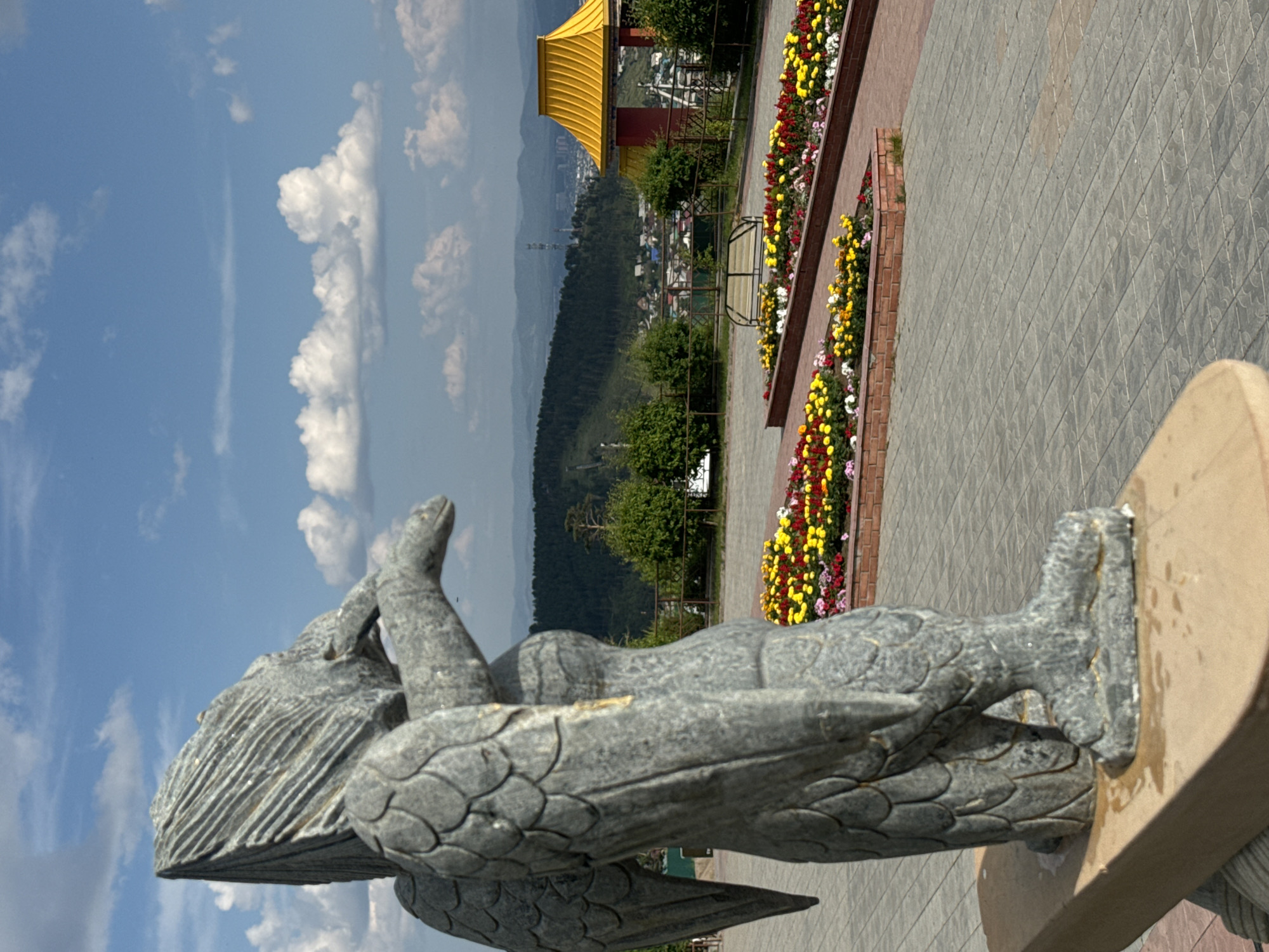 The image shows a stone sculpture of a winged creature in the foreground, positioned on a wooden base. The sculpture is made of gray stone and depicts a figure with detailed wings and a bird-like head. In the background, there is a well-maintained park area with paved walkways and flower beds planted with a variety of colorful flowers, including yellow, red, and pink blossoms. Further in the distance, a distinctive yellow-roofed pavilion with red accents is visible, suggesting a location of cultural or religious significance. Beyond the pavilion, a city landscape unfolds with buildings nestled among green hills and mountains under a bright blue sky with scattered white clouds. This scene is located at the "Wings of a Baikal Bird" sculpture park in Ulan-Ude, Russia, a popular viewpoint offering panoramic vistas of the city and its surroundings. There are no people visible in the image.