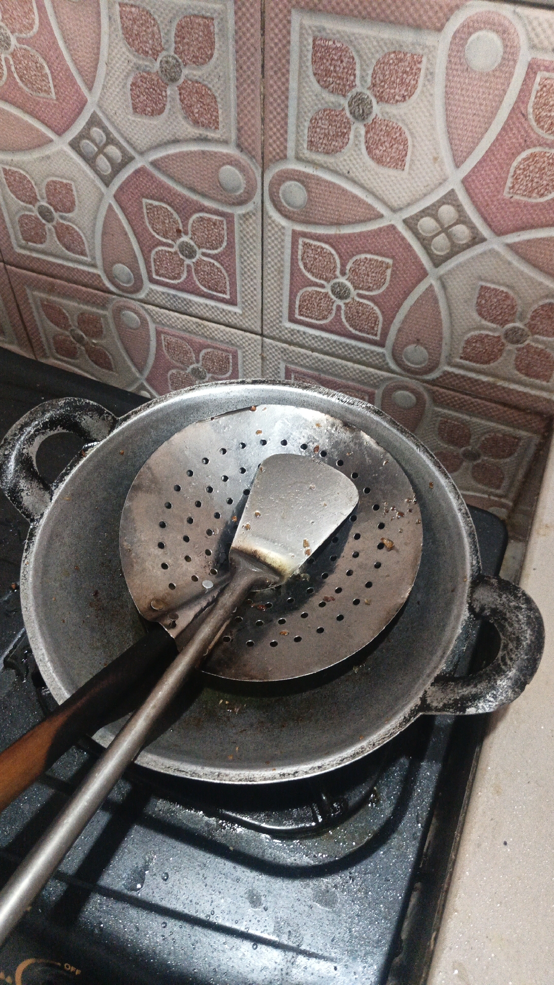 This image captures a close-up, overhead view of a used wok and cooking utensils placed on a stovetop. The wok, made of metal, is large and appears to have been recently used, with visible food remnants inside. A metal strainer with numerous holes is placed inside the wok, and a spatula rests on top of the strainer. The stovetop itself is black and shows signs of being used, with water or oil splatters on its surface. In the background, a wall is visible, tiled with a pink and white floral pattern. The scene suggests a domestic kitchen environment, likely in Kandangmas, Indonesia, where cooking has recently taken place. The lighting is adequate, allowing for clear visibility of the objects and their condition.