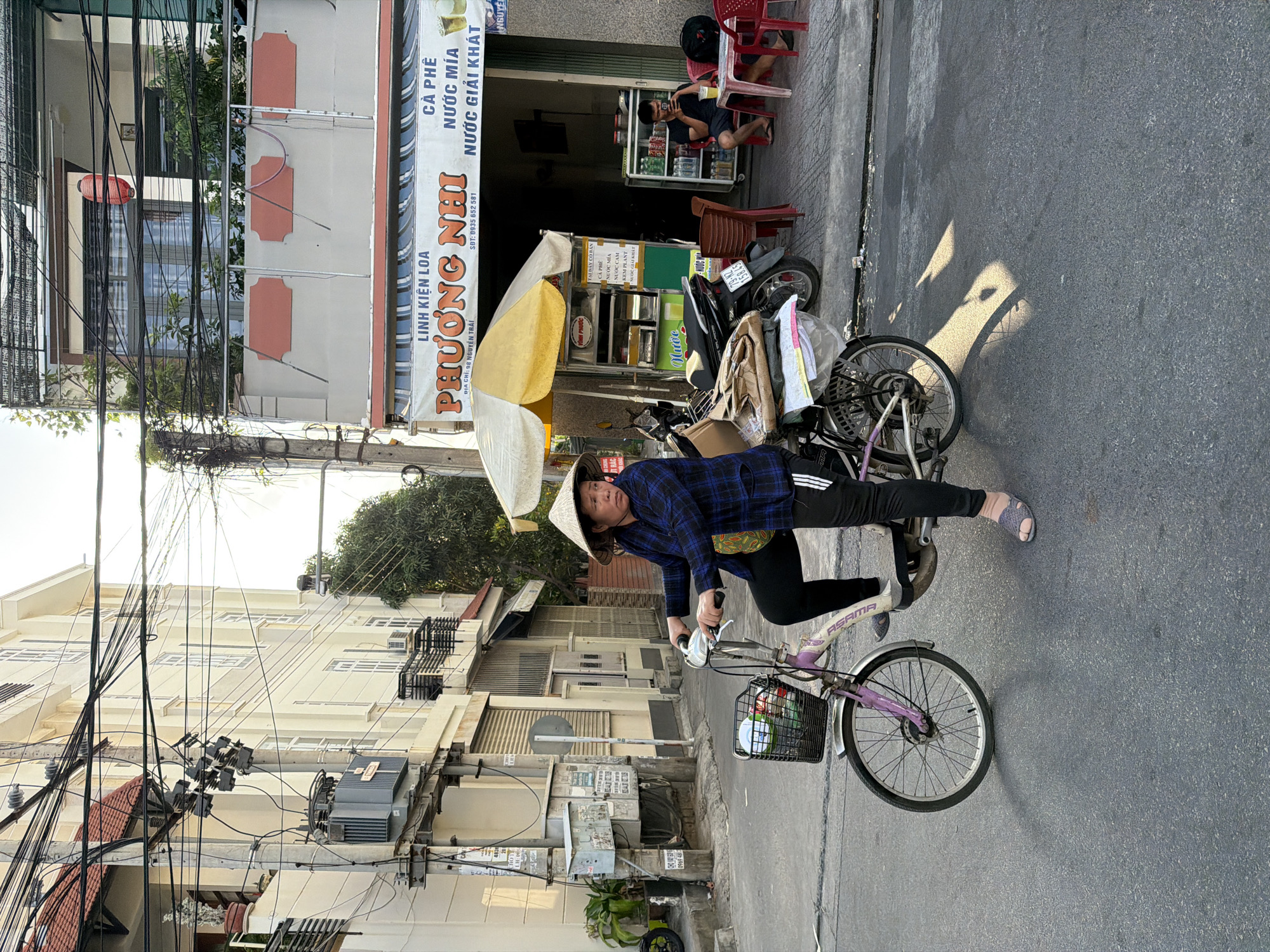 A woman wearing a conical hat and a blue checkered shirt is riding a purple bicycle on a paved street in Nha Trang, Vietnam. She is carrying a basket full of items on the front of her bicycle and has packages strapped to the back. The street is lined with buildings, including a shop with a sign that reads "PHUONG NHI LINH KIEN LOA" and offers "CA PHE NUOC MIA NUOC GIAI KHAT." A man is sitting at a table outside the shop, looking at his phone. The sun is casting long shadows on the street, indicating it is likely late afternoon. The scene appears to be a typical street view in a Vietnamese city, with its characteristic mix of residential and commercial buildings, busy streets, and street vendors.