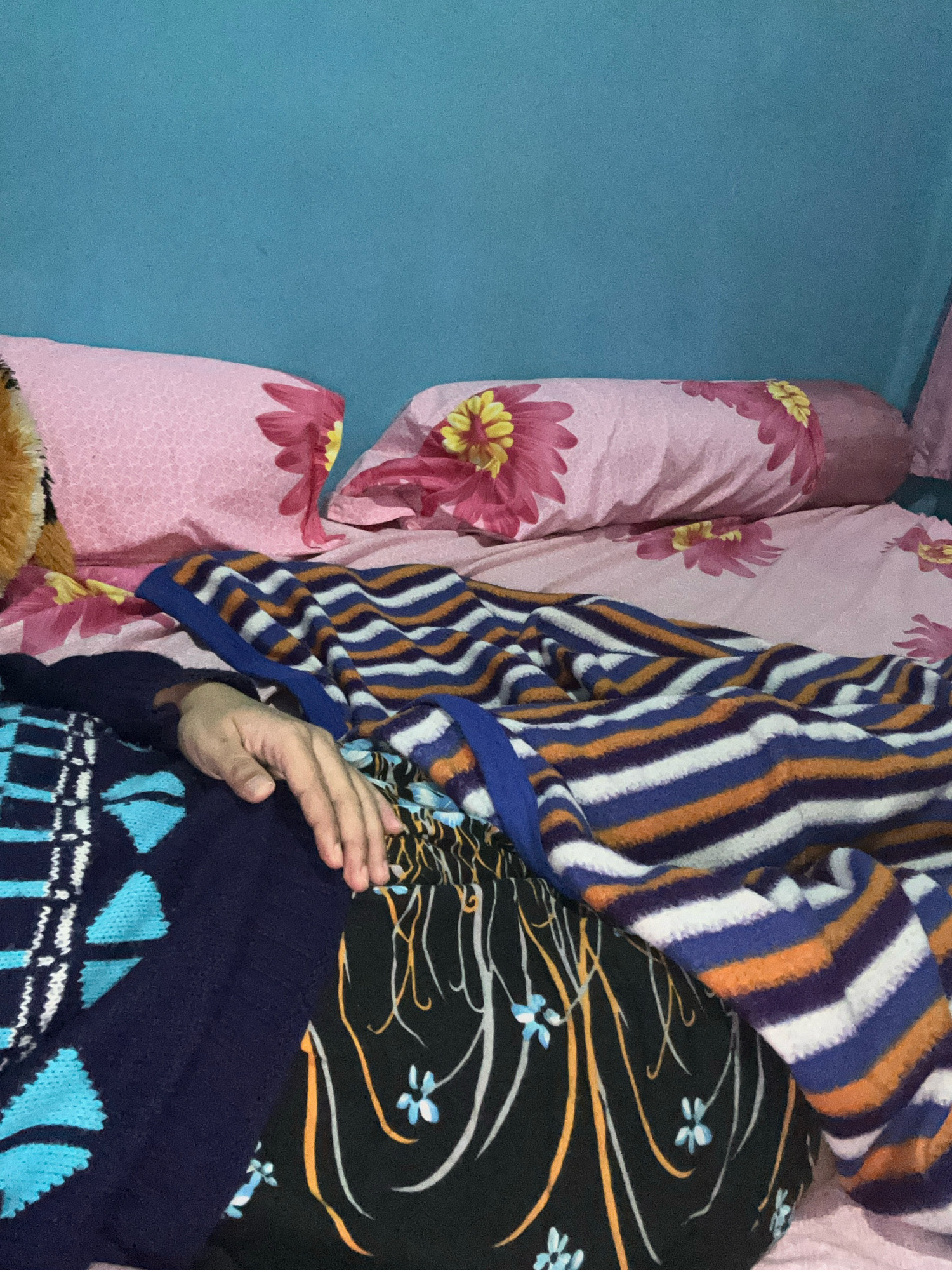 The image depicts a close-up view of a person lying on a bed. The bed is covered with a pink floral patterned sheet and a striped blanket with blue, orange, and white stripes. A person's hand is visible resting on top of the blanket, with fingers slightly curled. The background features a plain blue wall. The scene appears to be an indoor bedroom setting. There is no specific information in the image to suggest a location in Cihaurbeuti, Indonesia.