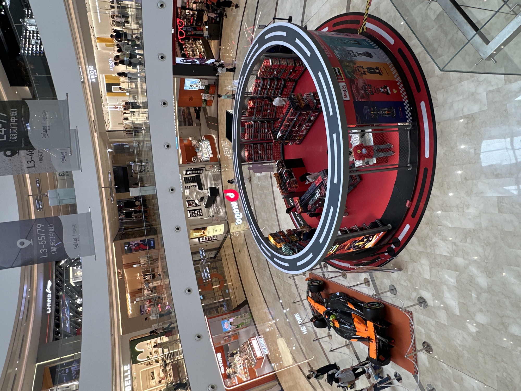 The image shows an overhead view of a shopping mall in Yuzhong District, China. The mall has multiple levels with various retail stores visible, including Ariose Tears, dzzit, Belle, Li-Ning, Neyone, and Hong Jiu. A circular display stands prominently in the mall's central area, featuring a racing-themed setup with toy race cars and branding for Maisto and Bburago. An orange and black toy Formula 1 car sits on a display platform with rope barriers and several people are milling about near the exhibit. The lighting is bright, consistent with an indoor retail environment. Signage visible includes "L3-40 (朝天门)", "L3-55/79 (朝天门中庭)", and references to CapitaLand.