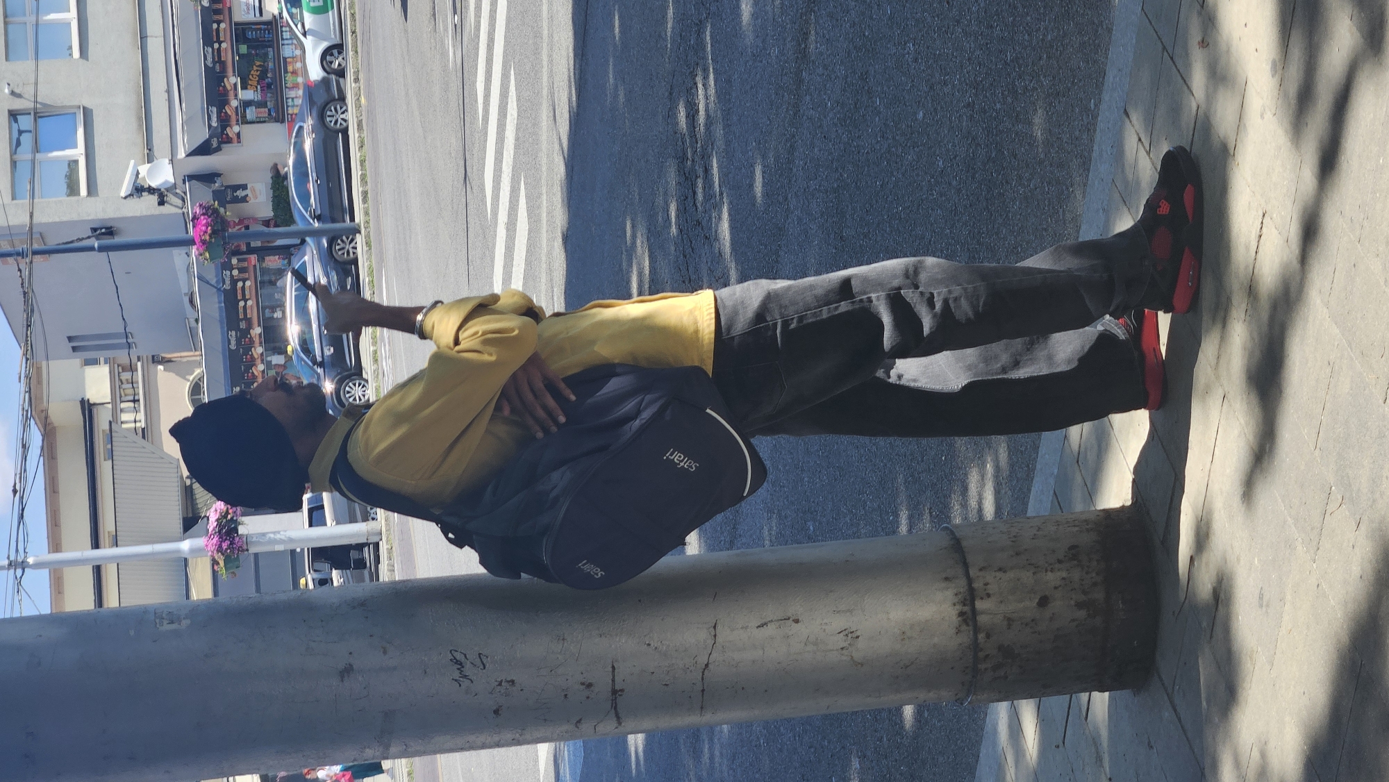A man wearing a black turban and a yellow shirt stands next to a metal pole, leaning his left shoulder against it. He has a dark backpack slung over his right shoulder and his left arm is crossed over his chest. In his right hand, he holds a cell phone up to his ear and appears to be speaking into it. He is wearing dark pants and red and black sneakers. The man is on a sidewalk in front of a street with cars and buildings in the background. The scene is lit by bright sunlight, casting shadows on the ground. The man's posture and the phone suggest he is in a conversation. The background includes a crosswalk, a building with windows, and a street sign that might indicate a location in Bratislava, Slovakia, though this is not definitively verifiable from the image alone.