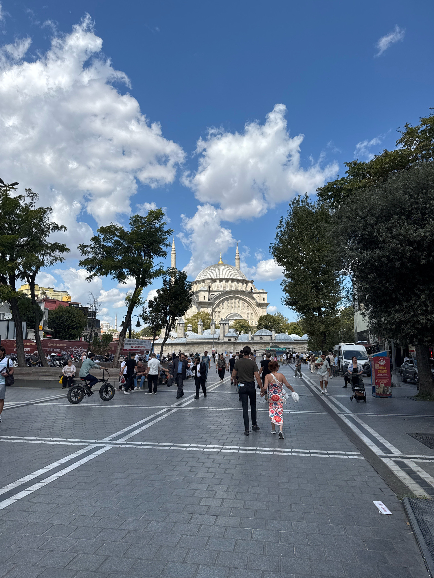 The image captures a vibrant street scene in Istanbul, Turkey, likely near the historic Sultanahmet Square, given the prominent view of a large mosque with multiple domes and a minaret. The sky is a bright blue with scattered white cumulus clouds. In the foreground, a paved street with white markings runs towards the mosque. Several people are walking along the street, dressed in casual summer attire. Some are individuals, while others are in pairs or small groups. A man is seen riding a black electric bicycle, moving away from the camera. Trees line the sides of the street, and buildings are visible in the background, some with traditional Ottoman architecture. The atmosphere appears lively and bustling, with a sense of everyday urban life.