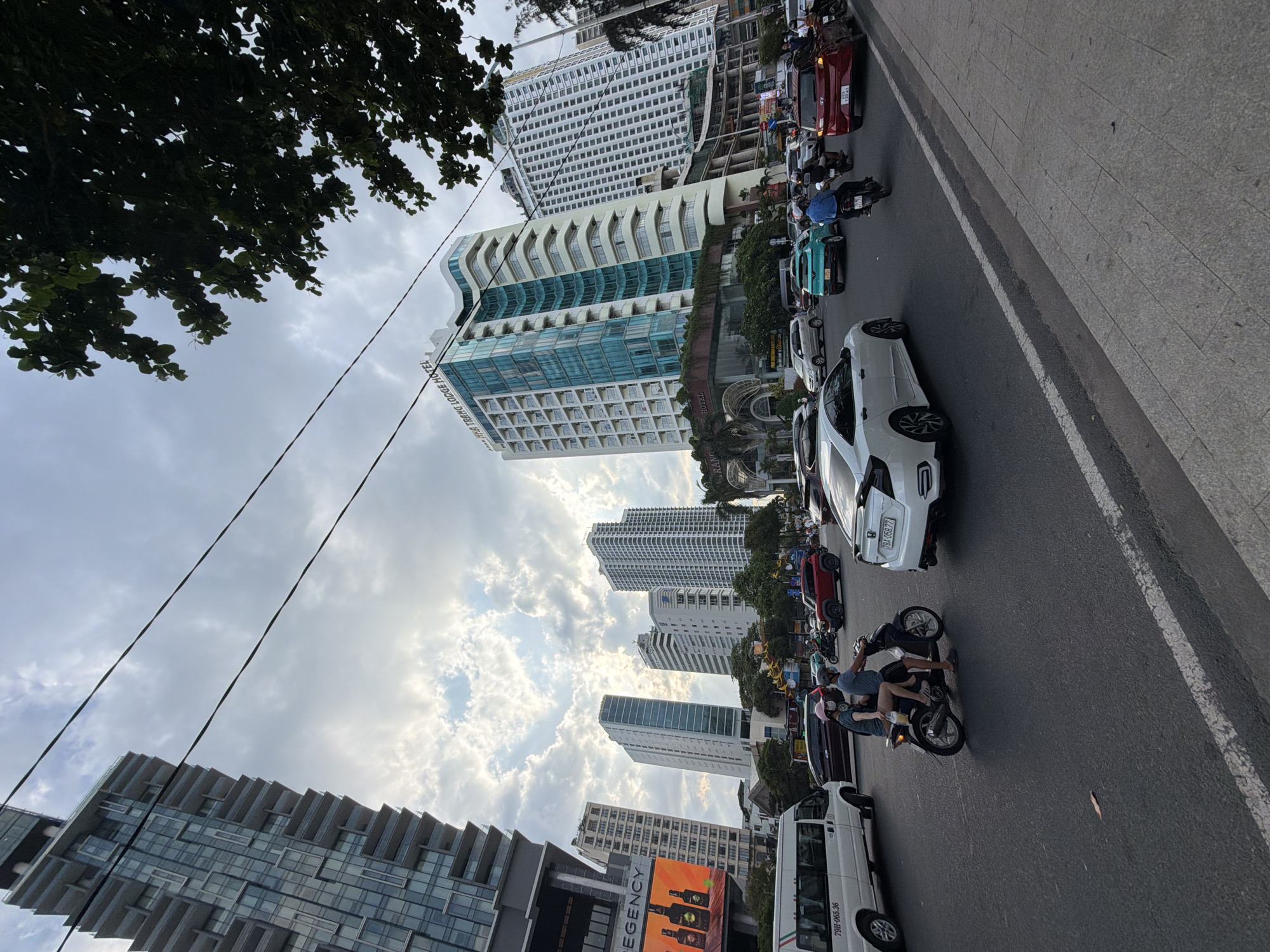 This image captures a bustling urban street scene in Nha Trang, Vietnam, with modern high-rise buildings dominating the skyline. The sky, visible between dense tree foliage on the left and towering structures, is partly cloudy with patches of bright light, suggesting a daytime, possibly late afternoon, setting.
The foreground features a multi-lane asphalt road with a wide paved sidewalk to the right. Traffic includes numerous cars and motorbikes. A prominent white car (possibly a Honda Civic) is seen in the foreground, with its license plate "79H-063.36" visible. Further down the road, a red car with license plate "79A-193.41" is in a queue. Multiple motorbikes with riders, some wearing helmets, navigate the road. One person on a motorbike is wearing a blue rain poncho. A white van with license plate "79H-065.36" is also visible.
Several tall, contemporary buildings define the urban environment. A distinctive building with a wavy, glass-heavy facade is identified by the vertical text "THE TRUNG LODGE HOTEL" on its side. Another large building behind it features a grid pattern of windows. To the far left, part of another modern high-rise with stepped architectural details is visible. A billboard or sign at the base of this building displays the text "REGENCY" and images of bottles. Power lines are visible stretching across the sky.