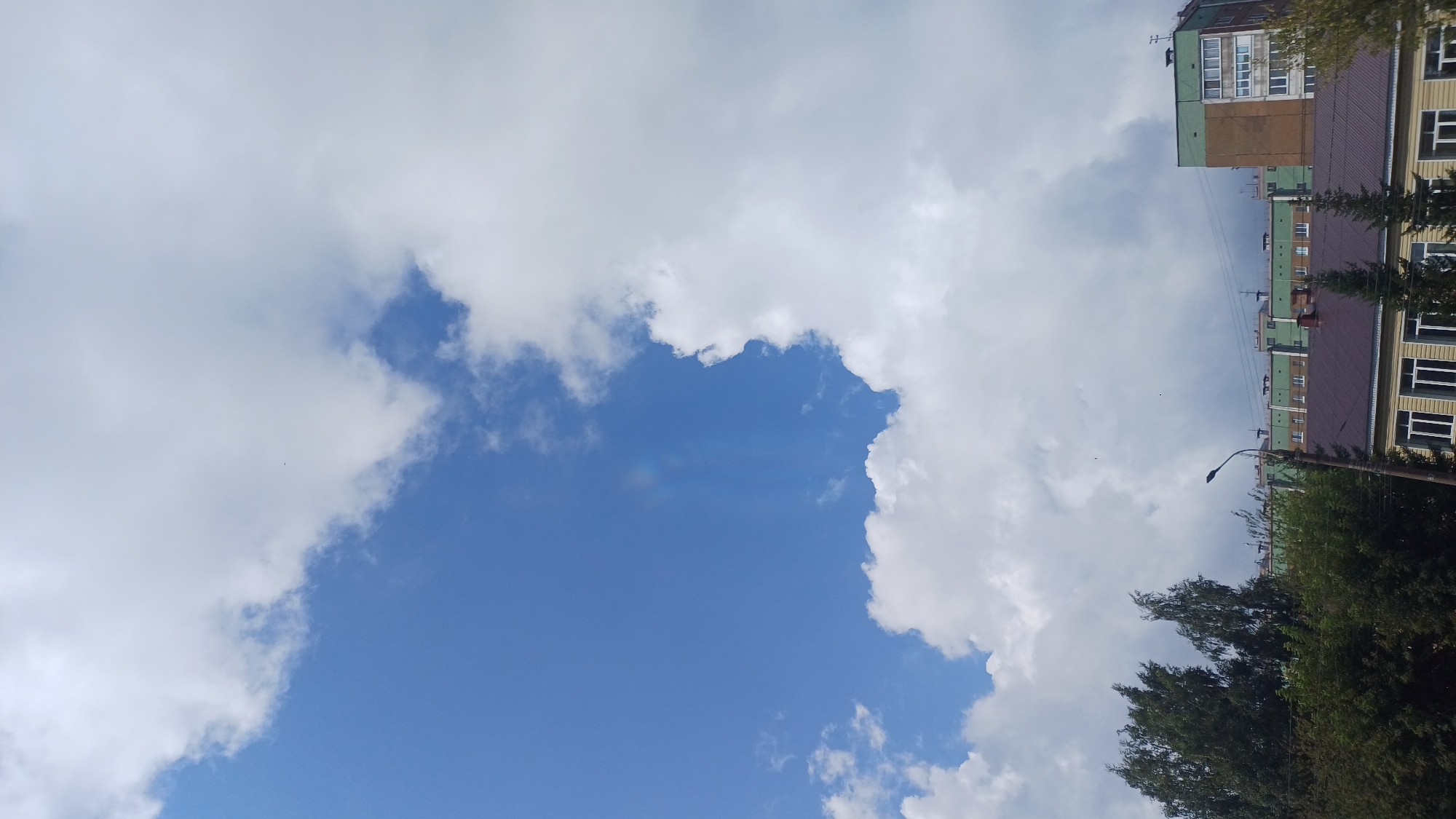 The image shows a sky with scattered clouds and a faint rainbow. Below the sky, there are several buildings, some with green facades, and trees. A street lamp is visible in the foreground. The scene is set in Izhevsk, Russia.