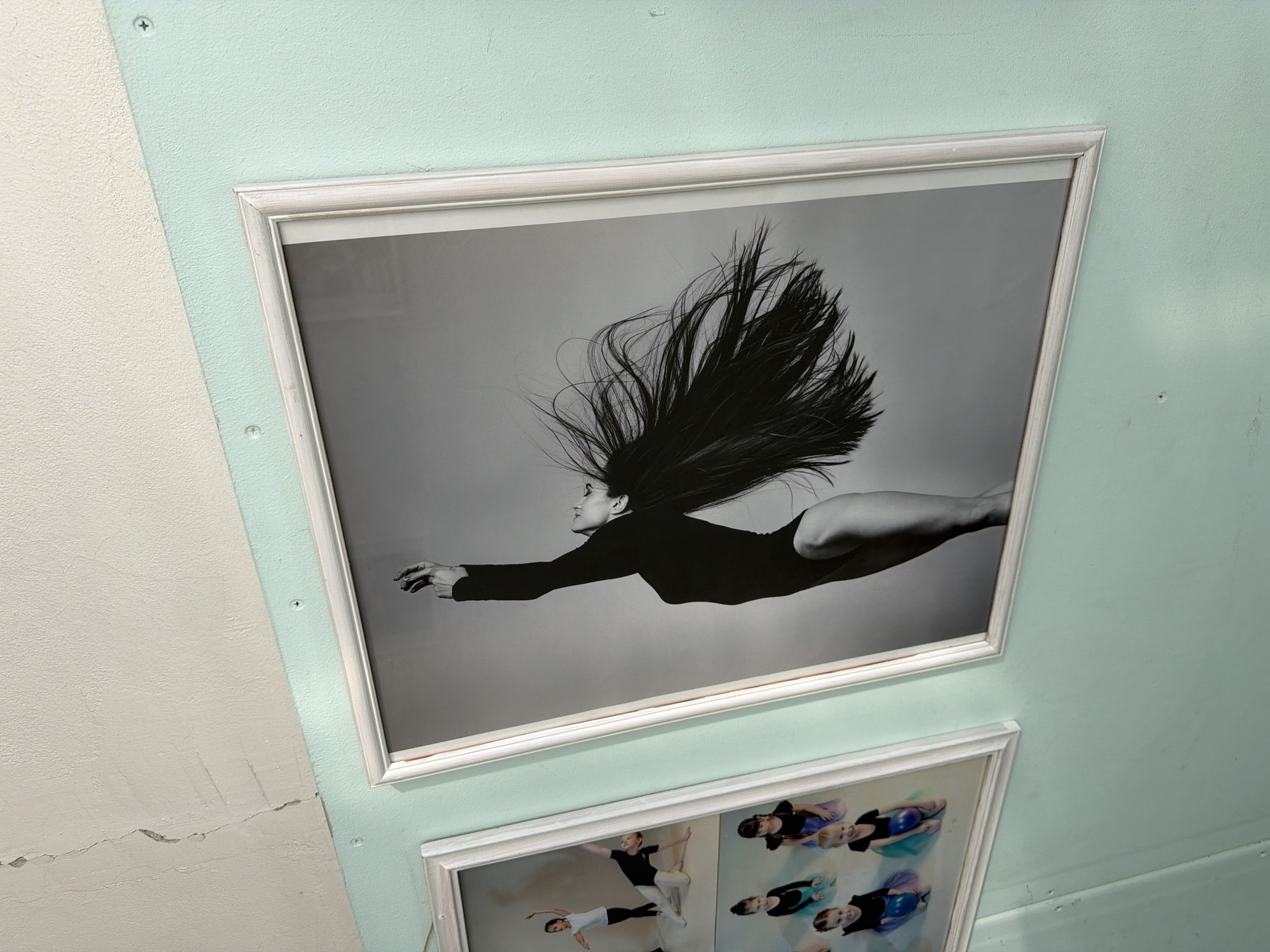 A framed black-and-white photo of a woman in a bodysuit with her hair flowing is hung on a pale green wall in a room. Below it is another framed photo of a group of children. The wall appears to be in a building, but its location within the city of Saint Petersburg, Russia, cannot be determined from this image alone.