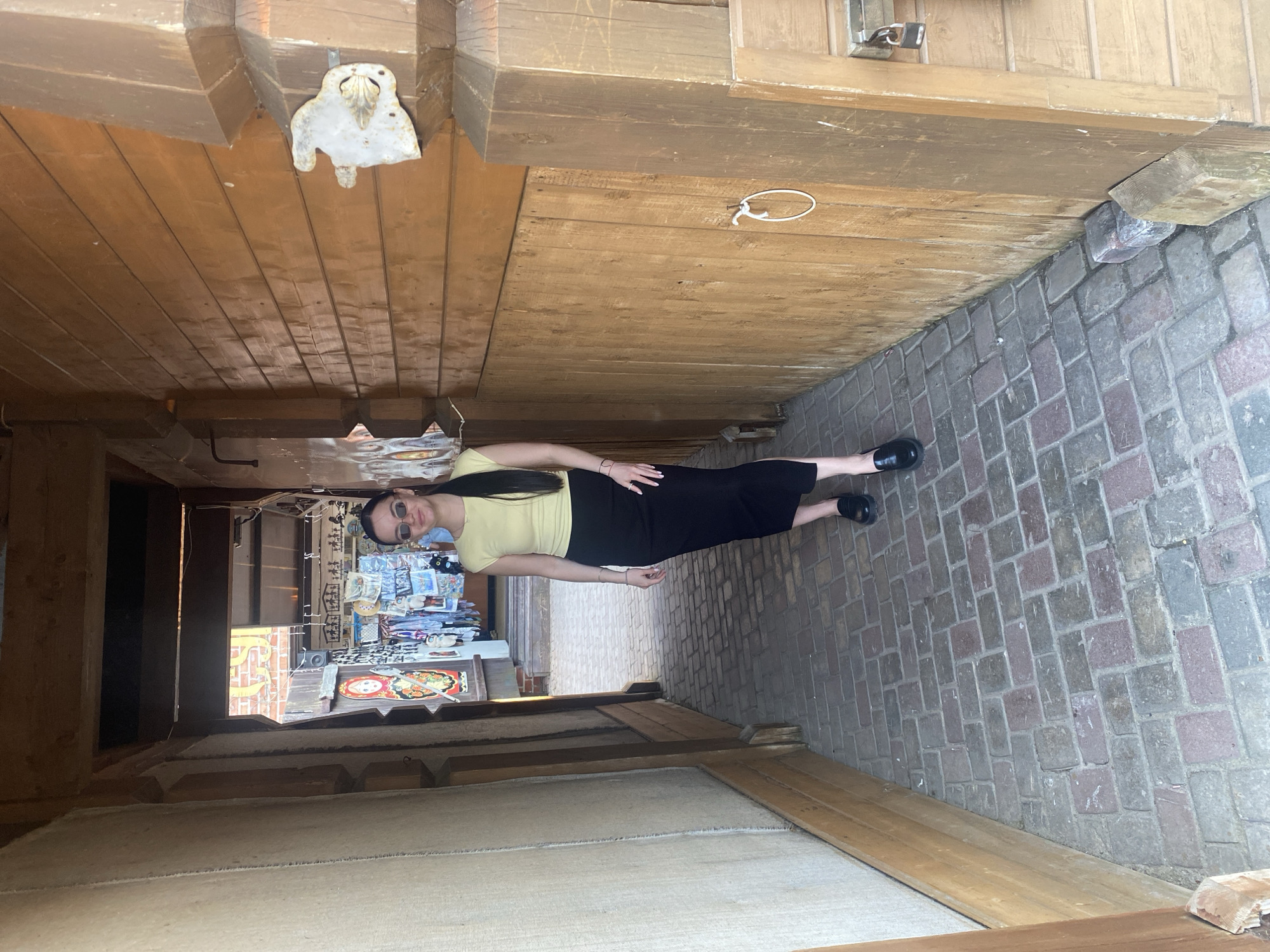 A woman wearing sunglasses, a yellow shirt, and a black skirt is standing in a covered walkway or alleyway in the city of Sviyazhsk, Russia. The walkway has a wooden ceiling and the floor is paved with brick. A shop with souvenirs is visible in the background.