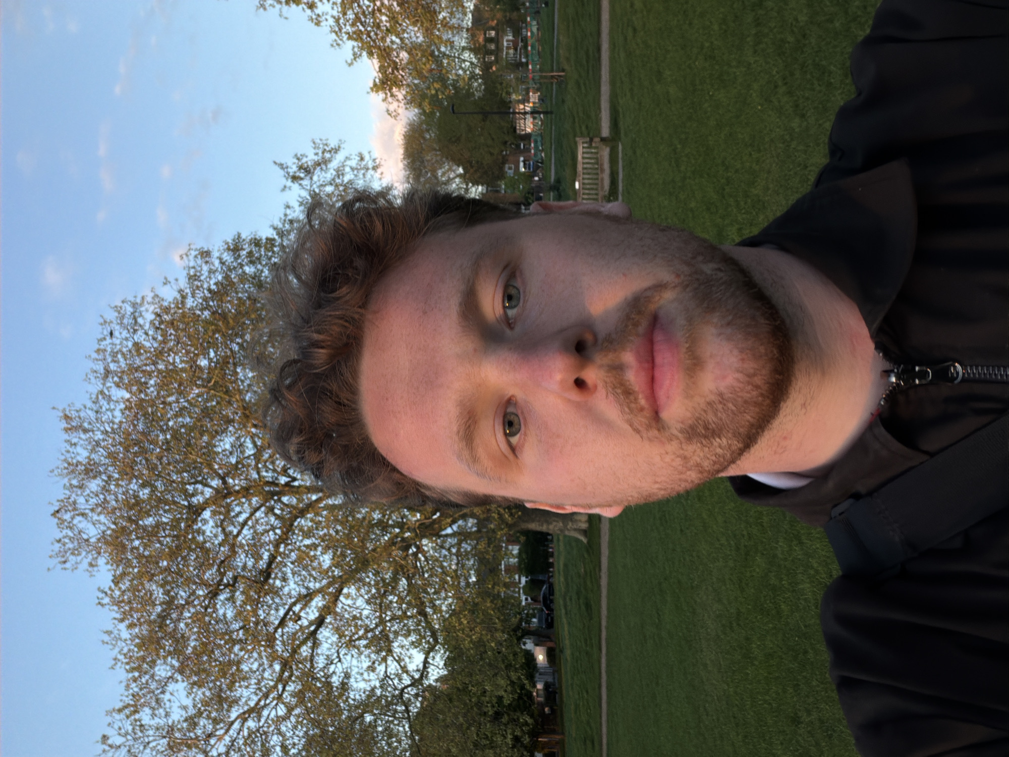 A selfie-style image features a fair-skinned male with brown hair and a beard. The photo appears to be taken in a park or green space, given the presence of green grass, a large tree with visible branches, and a blue sky with light clouds in the background. The man is wearing a dark-colored jacket. In the background, there are glimpses of urban features, including buildings and street furniture, suggesting a location in a city. Given the style of buildings and the context, the city is likely London, United Kingdom.