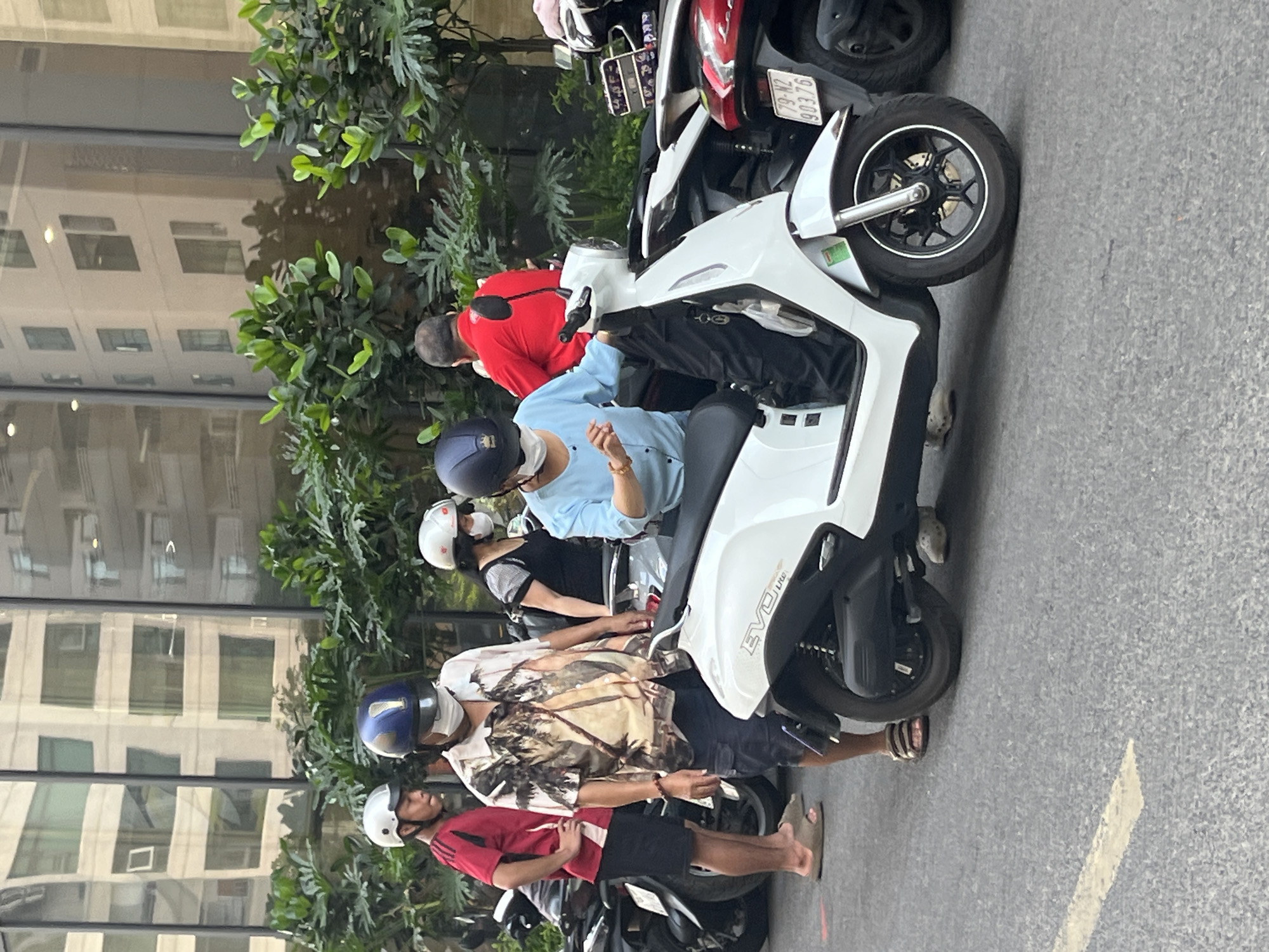 In Bac Nha Trang Ward, Vietnam, an outdoor scene unfolds with several individuals and their scooters. On the left, a young man in a red t-shirt and black shorts stands beside a motorcycle. To his right, a man wearing a patterned shirt, dark shorts, and a blue helmet is seated on a white scooter. A woman in a light blue shirt and a white helmet is also on the white scooter, positioned in front of the man. Behind them, another woman in a black top and a white helmet stands near a parked scooter. Further to the right, a man in a red shirt is visible. The background features lush green foliage and the reflection of a modern building on a glass facade. The scooters are parked on a grey asphalt surface with a white line marking.