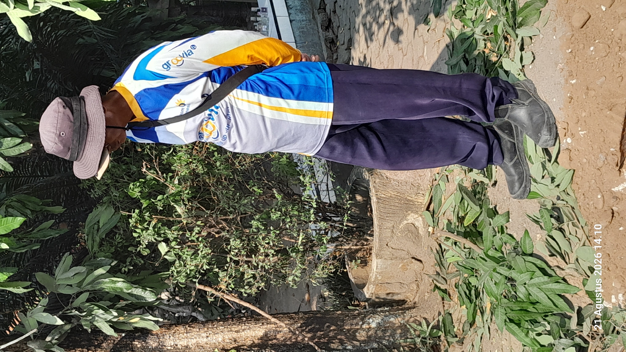 A man wearing a grey wide-brimmed hat, a blue and white long-sleeved shirt with yellow accents, and navy blue pants stands in an outdoor environment. He has a black strap across his chest. The man is looking down and to his right. The ground around him is covered in fallen green leaves and branches. In the background, there is a small green bush in a concrete planter, a tree trunk, and foliage. To the far right, a white structure with bottles on it is visible. The date stamp "21 Agustus 2026 14.10" is present in the lower left corner, suggesting this image was taken in Takeran, Indonesia.
