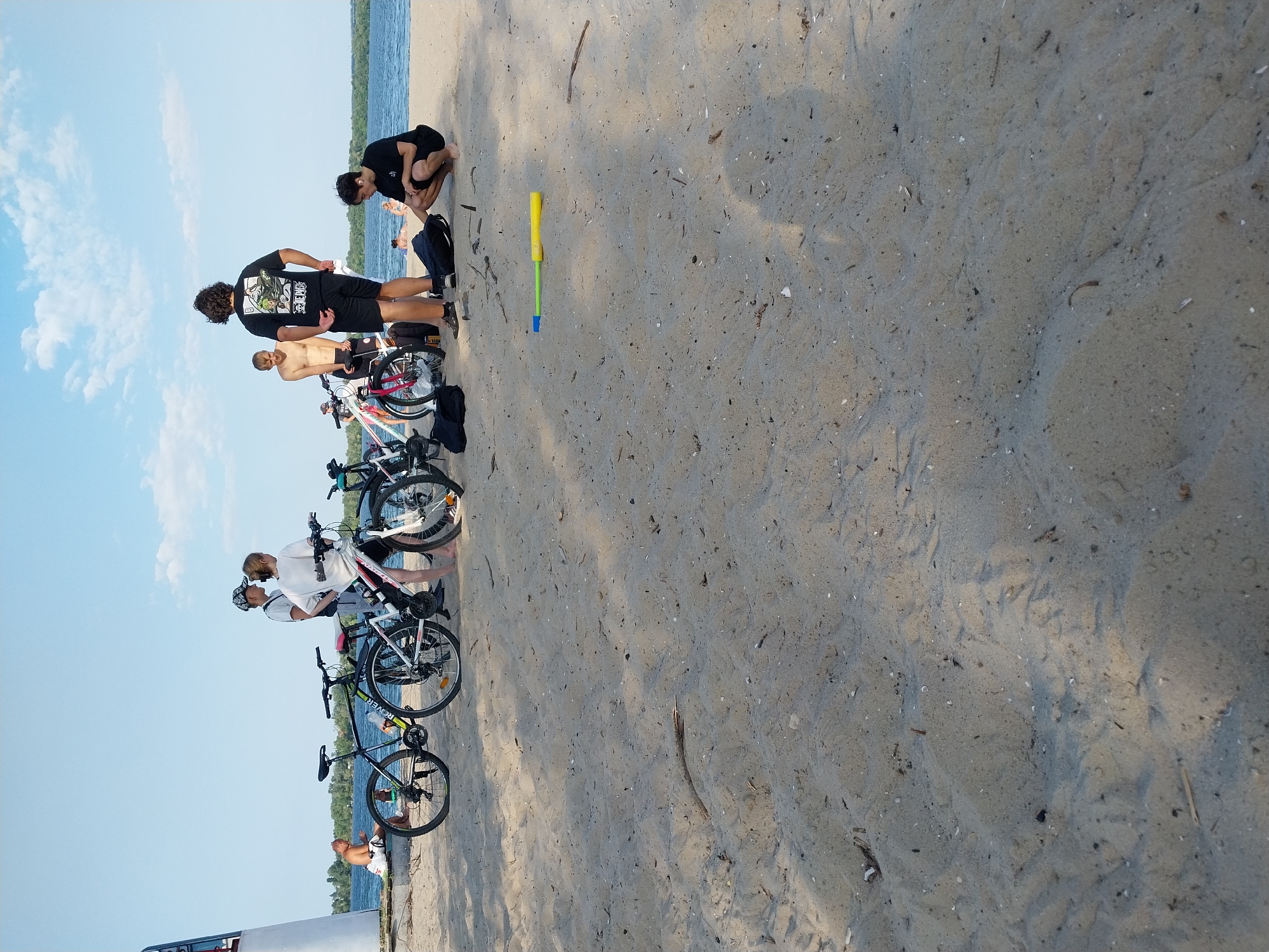 A group of young people are gathered on a sandy beach next to a body of water. Several bicycles are parked nearby. One person is sitting on a bench, facing away from the camera. Another person is squatting on the sand, looking at something in front of them. The sky is blue with scattered clouds. The overall scene appears to be a recreational outing on a sunny day.
