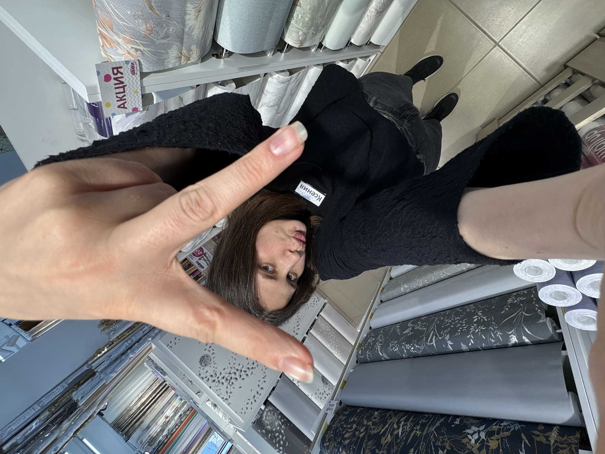 A woman is taking a selfie inside a retail store specializing in wallpaper and window treatments. The woman has shoulder-length dark hair and is wearing a black textured top with a name tag that reads "Ксения." She is making a "peace sign" with her left hand, which is close to the camera.
The woman is standing in an aisle of the store, which is filled with shelves of wallpaper rolls and stacks of fabric samples. A promotional sign with the word "АКЦИЯ" is visible. The woman is wearing dark jeans and black shoes. The floor tiles are a light beige color. The setting appears to be a store in Chaikovsky, Russia.