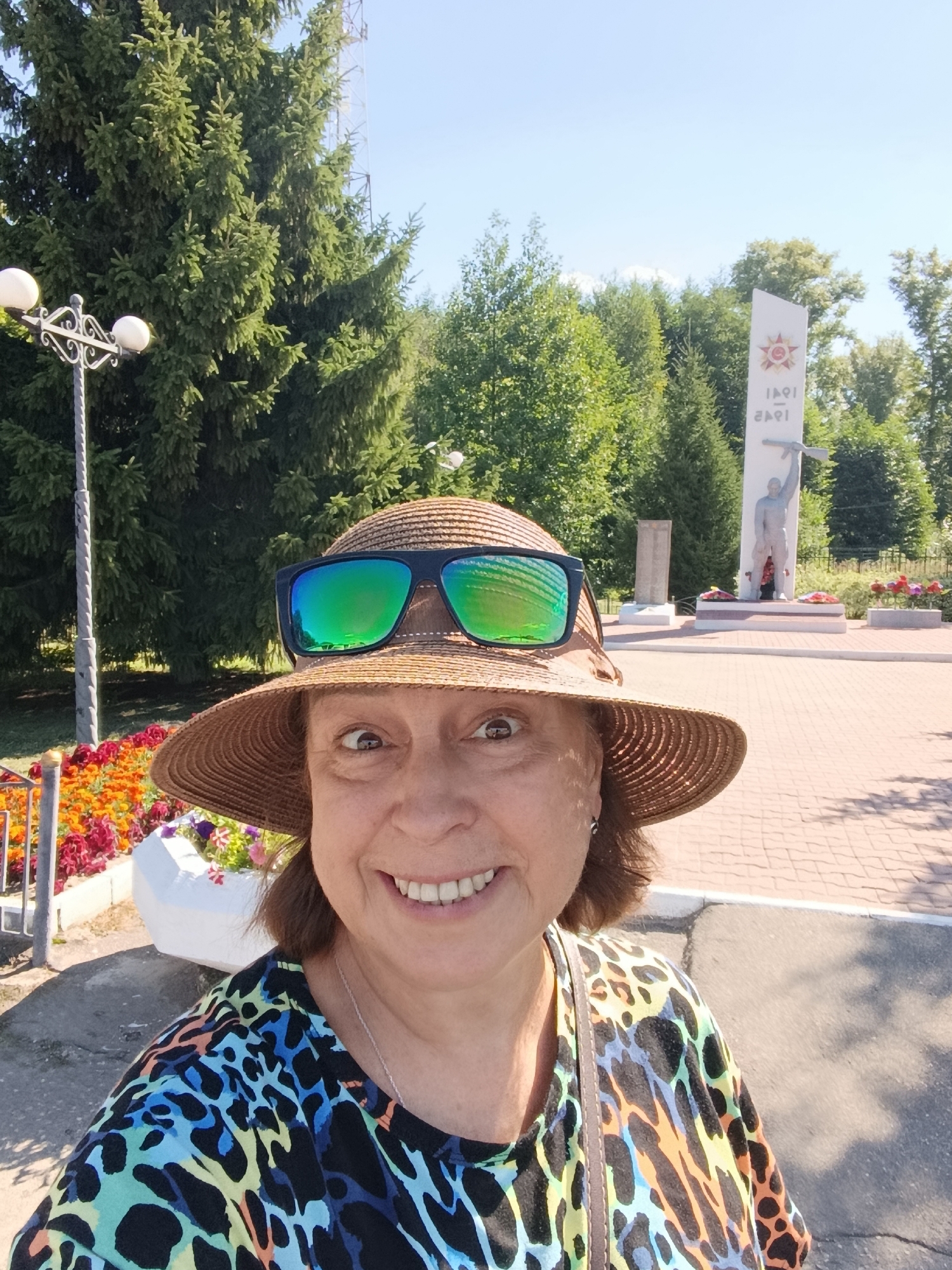 A woman wearing a straw hat and sunglasses takes a selfie in front of a memorial. She has brown hair and is smiling. Her shirt has a colorful leopard-print pattern. In the background, a tall white monument with a red star and the dates "1941-1945" is visible, along with a statue of a soldier holding a rifle. There are trees and flowers surrounding the monument. The ground is paved with brick-like stones. The scene appears to be a sunny day in the city of Vereya, Russia, likely at a war memorial.