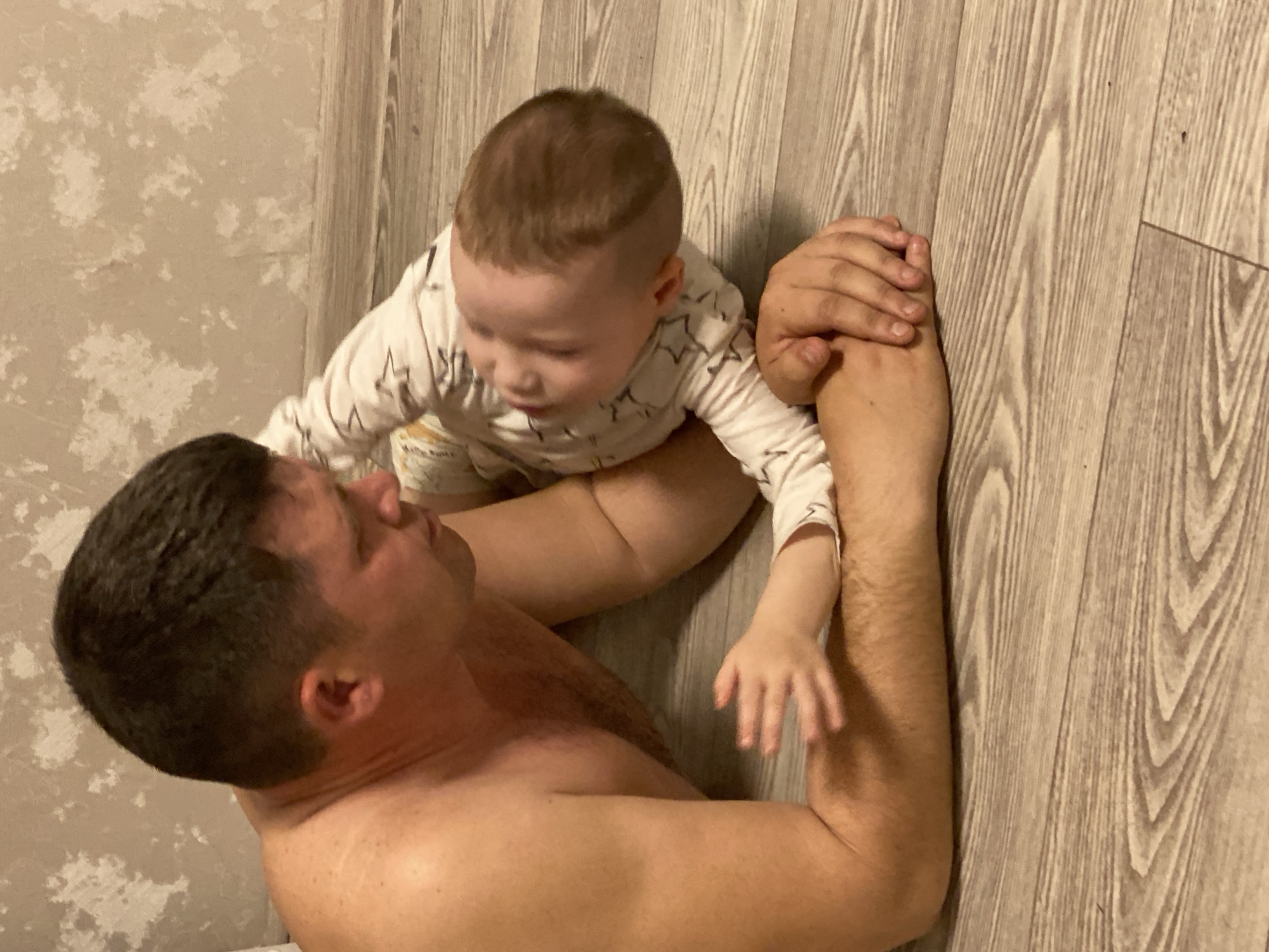 A Caucasian man is lying on a light wood-patterned floor in an indoor setting, holding a baby on his body. The man is shirtless with dark hair, and the baby is wearing a white pajama top with black star patterns. The baby’s hand rests on the man's arm. The setting includes a wall with a textured surface. The location is Sochi, Russia.