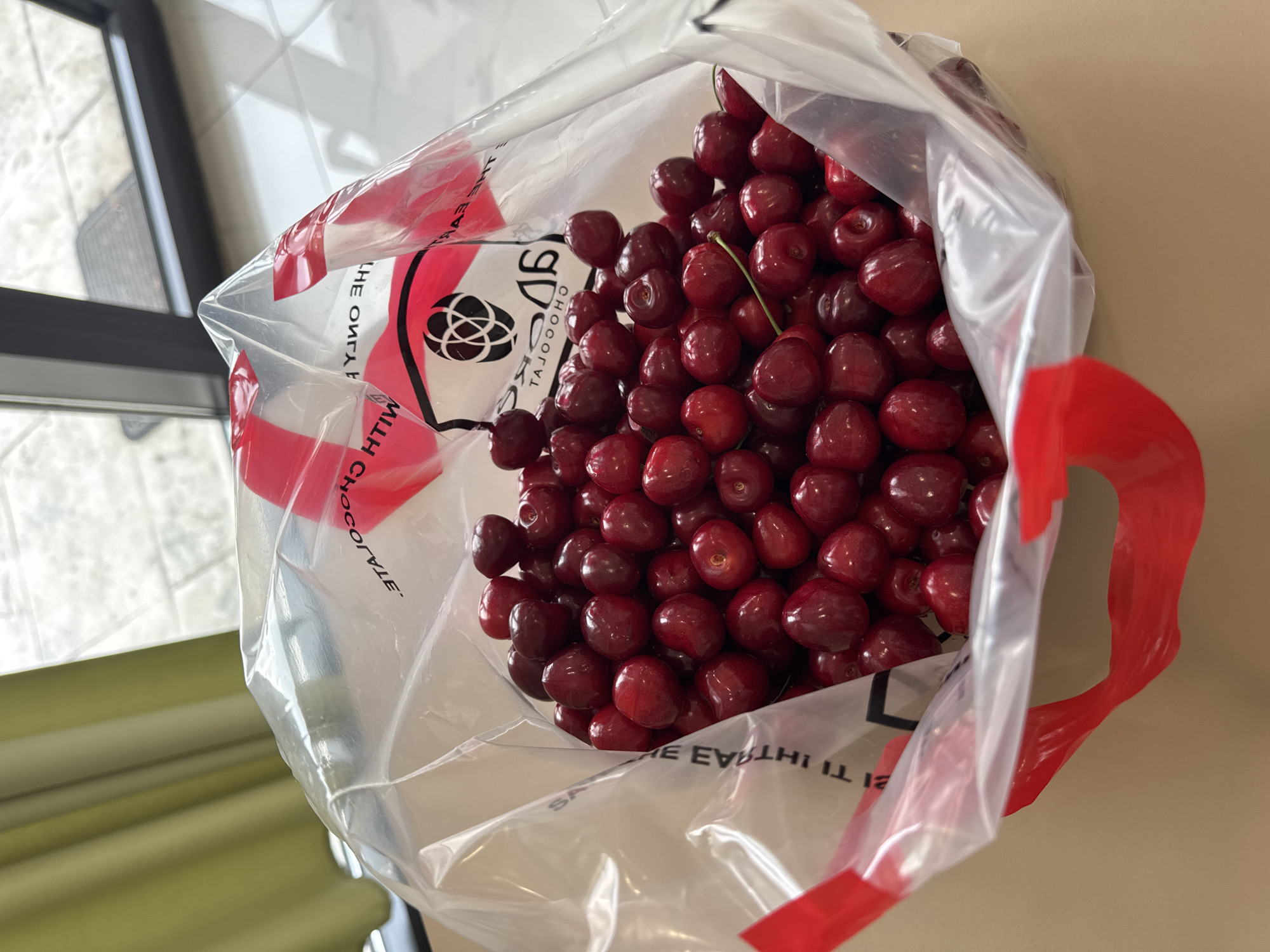 The image shows a bag of red cherries. The bag is clear plastic with red accents and writing in an unknown language. The cherries fill the bag, obscuring much of the text. The background is a light beige surface and a window with a view of what appears to be an outdoor area. There is no evidence in the image to suggest a location within the city of Podgorica, Montenegro. No people are visible.