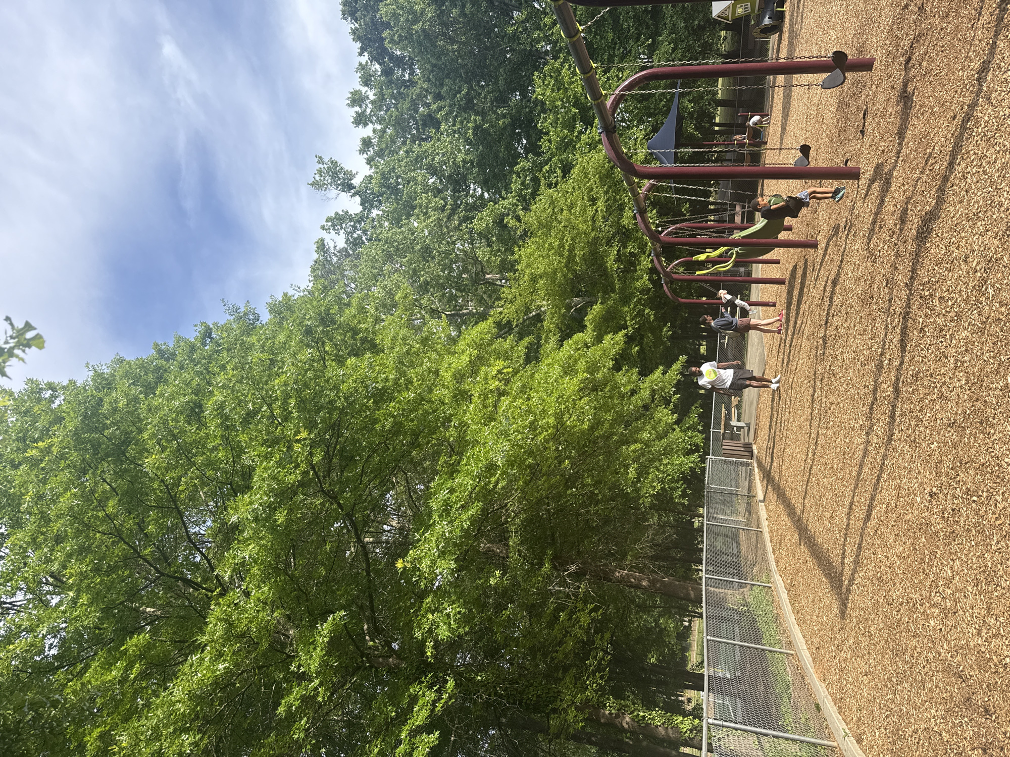 The image shows a playground in Mountain Park, United States of America. The playground has a wood chip ground cover and a metal fence bordering it. A swing set with red frames stands on the right side of the image. Several people are visible, including a child lying on a slide and other individuals standing or walking near the swings. Dense green trees dominate the left side and background, with patches of blue sky visible above.