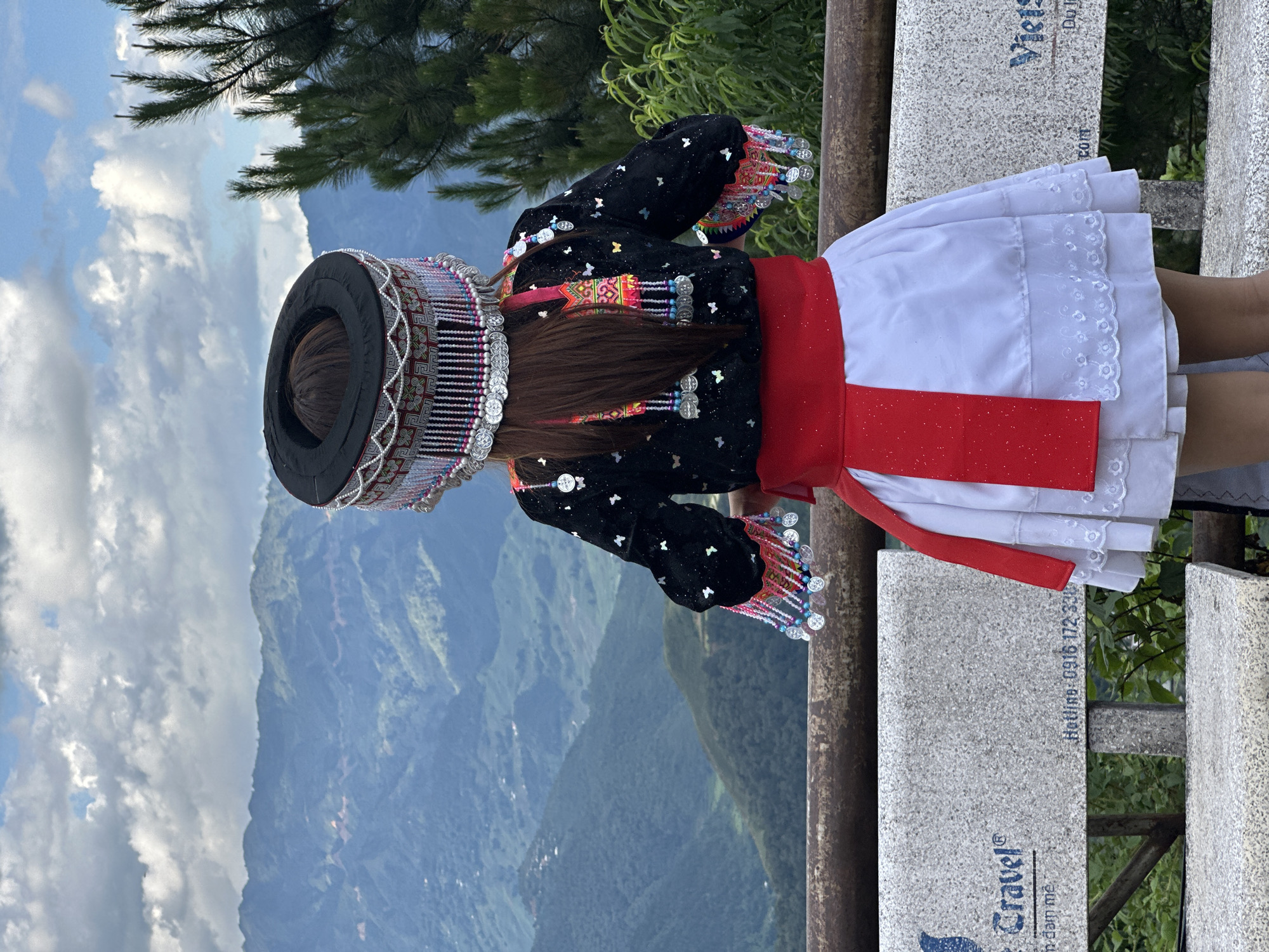 A person, viewed from the back, stands on a viewing platform overlooking a vast mountainous landscape in Tu Le, Vietnam. The individual wears traditional attire consisting of an elaborate black circular headpiece adorned with intricate silver and white beadwork, colorful small beads (pink, blue), and multiple dangling silver coin-like discs. Long brown hair is visible beneath the headpiece. The person wears a black long-sleeved top embroidered with colorful patterns (pink, yellow, green) on the upper back and cuffs, embellished with small silver butterfly-shaped appliques and additional dangling silver discs from the sleeves and back. A wide, sparkly red sash is tied around the waist, above a white tiered skirt featuring white floral embroidery on its lowest tier. The person stands behind a weathered wooden railing. In the background, rolling green mountains recede into the distance under a sky with scattered white and grey clouds. Sparse green foliage is visible on the right side of the frame. Below the railing, a grey granite bench is visible, displaying partial text including "Travel®" with a blue logo, "n dam me," "Hotline: 0916 172 338," and "Viets" alongside "Du l."