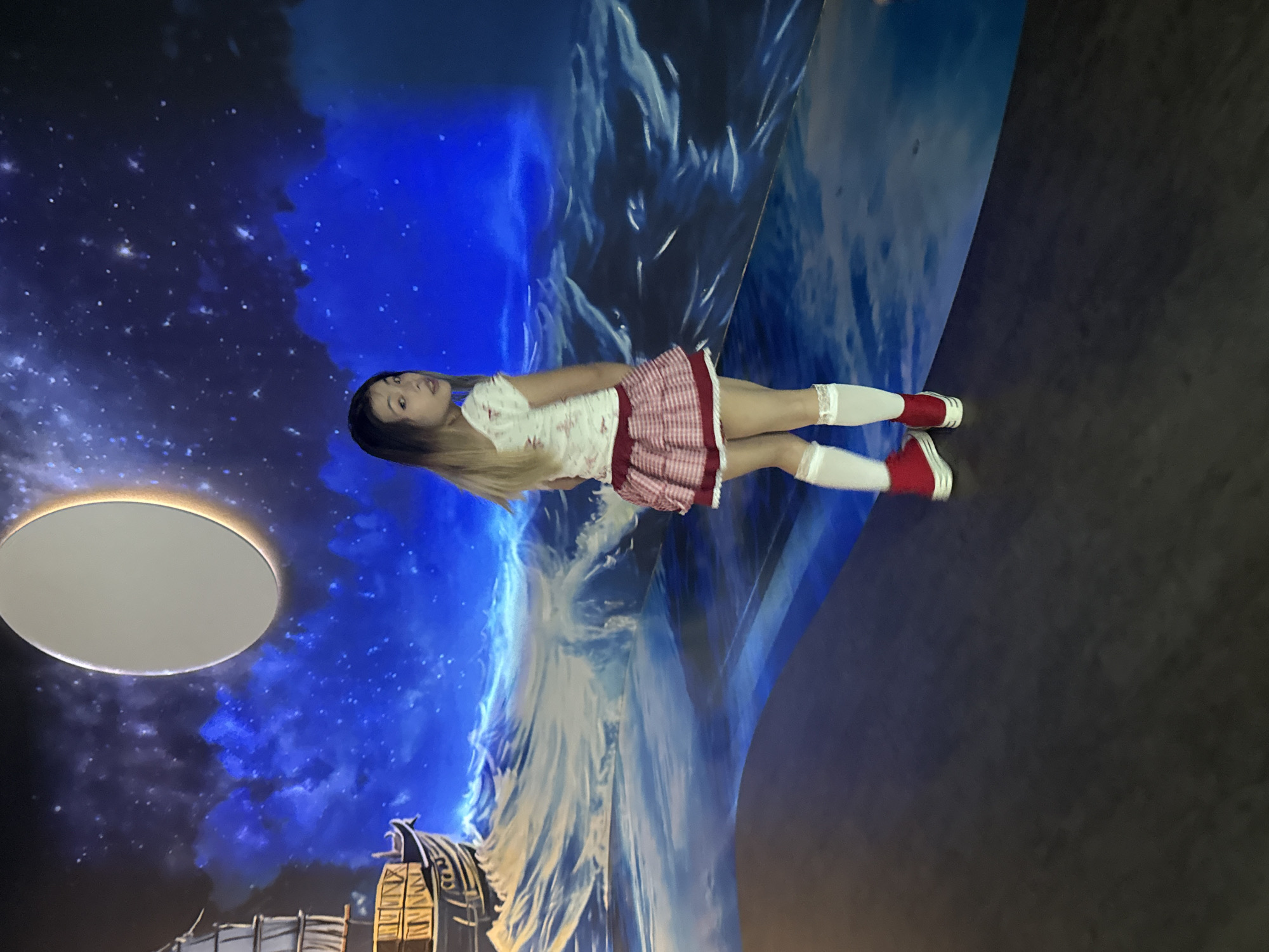 The image captures a full-length shot of a young woman standing in what appears to be an immersive art installation. She has long blonde hair, fair skin, and is wearing a short, patterned red and white skirt, a white top, white knee-high socks, and red sneakers.
The background is a dynamic display of colors and patterns. There's a blend of dark blues suggesting a night sky, light blue and white that resemble a mountain landscape, and some structural elements towards the bottom that could be part of the art installation. To the left, there is a large, circular, illuminated panel, possibly a light fixture or another element of the installation.
While the location within the city of Yuzhong District, China, isn't explicitly visible from the image, it can be inferred that this image was taken in an art gallery or immersive art experience space, such as the Chongqing Yuzhong Peninsula Cloud Art Gallery. The scene suggests an environment designed to create a unique visual and sensory experience.