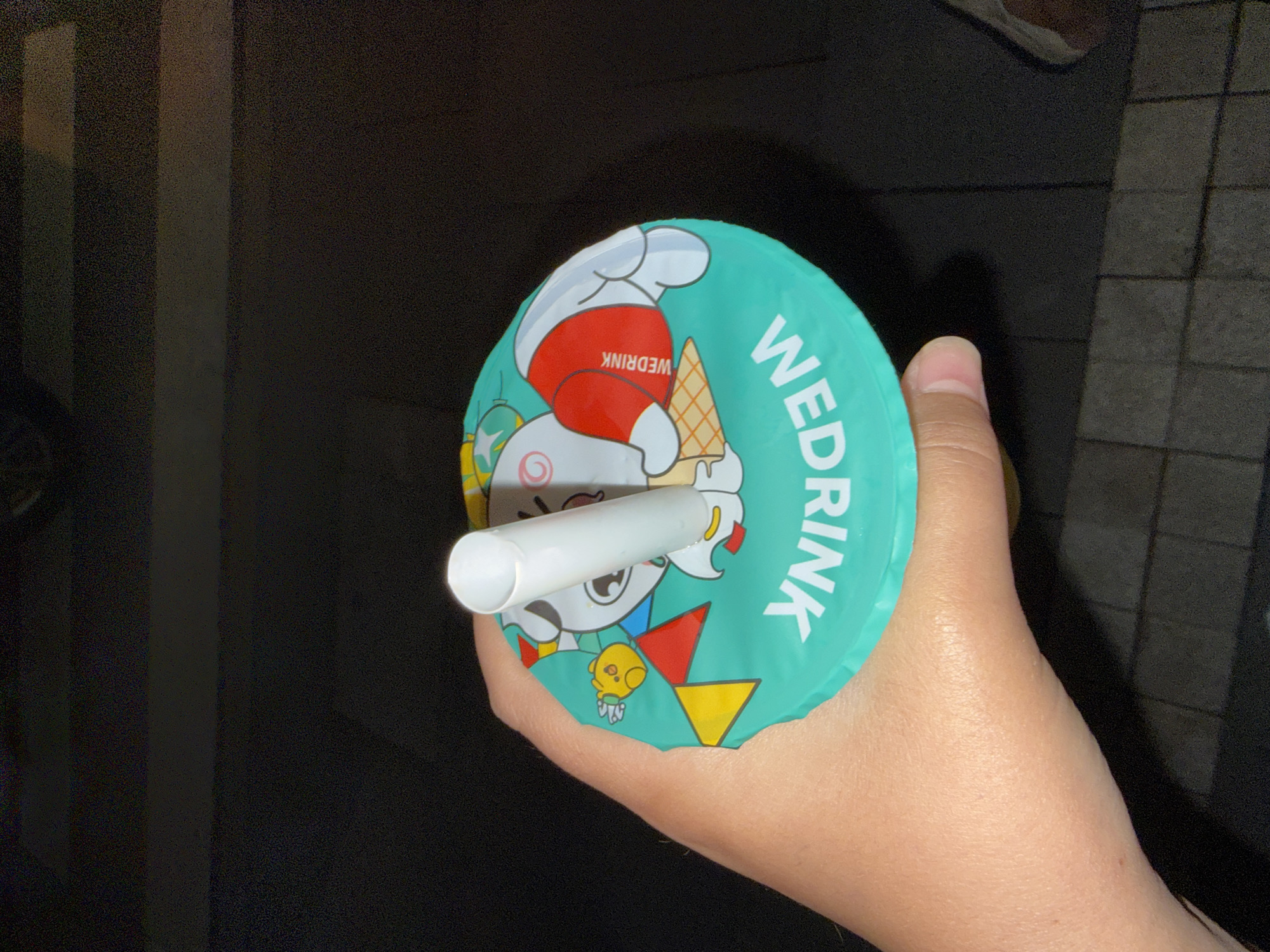 An overhead shot shows a hand holding a teal-colored cup lid with a cartoon character and "WE DRINK" logo. A white straw is inserted through the lid. The background is dark, with glimpses of what appears to be paving stones. No specific location in Astana, Kazakhstan is identifiable from the image.