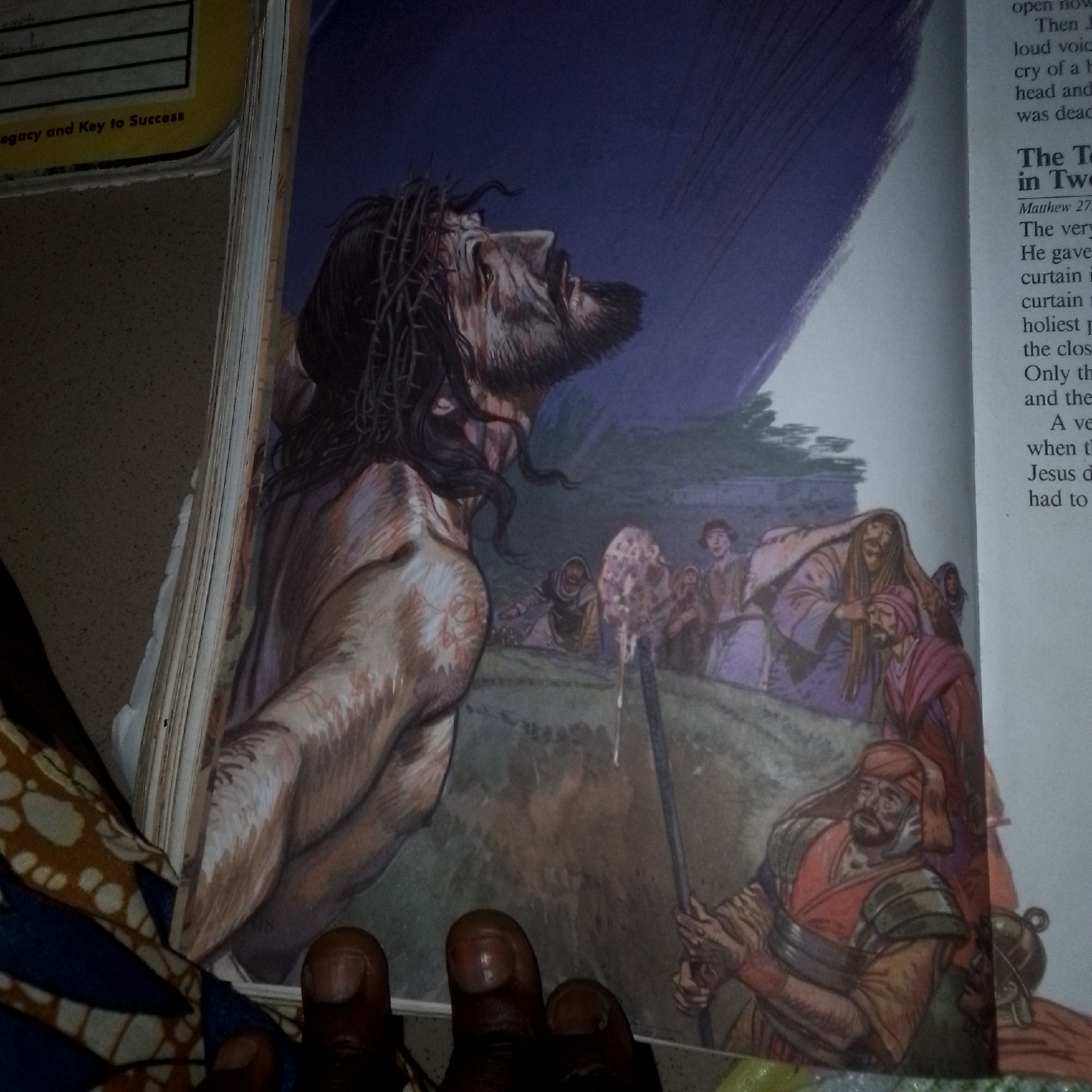 The image depicts an open book with an illustration of Jesus wearing a crown of thorns, with his head bowed and looking upwards. He is bloody and appears to be in distress. Behind him, a group of people are gathered, some appearing to be observing the scene. The background is a dark, swirling purple, possibly representing the sky. On the right side of the image, text from the book is visible, with headings like "The T" and "in Two". The illustration style is realistic and somewhat dramatic. The bottom of the image shows a portion of a patterned fabric and several fingers, suggesting the book is being held or is resting on a surface with this fabric. There is no indication of any specific city or location.