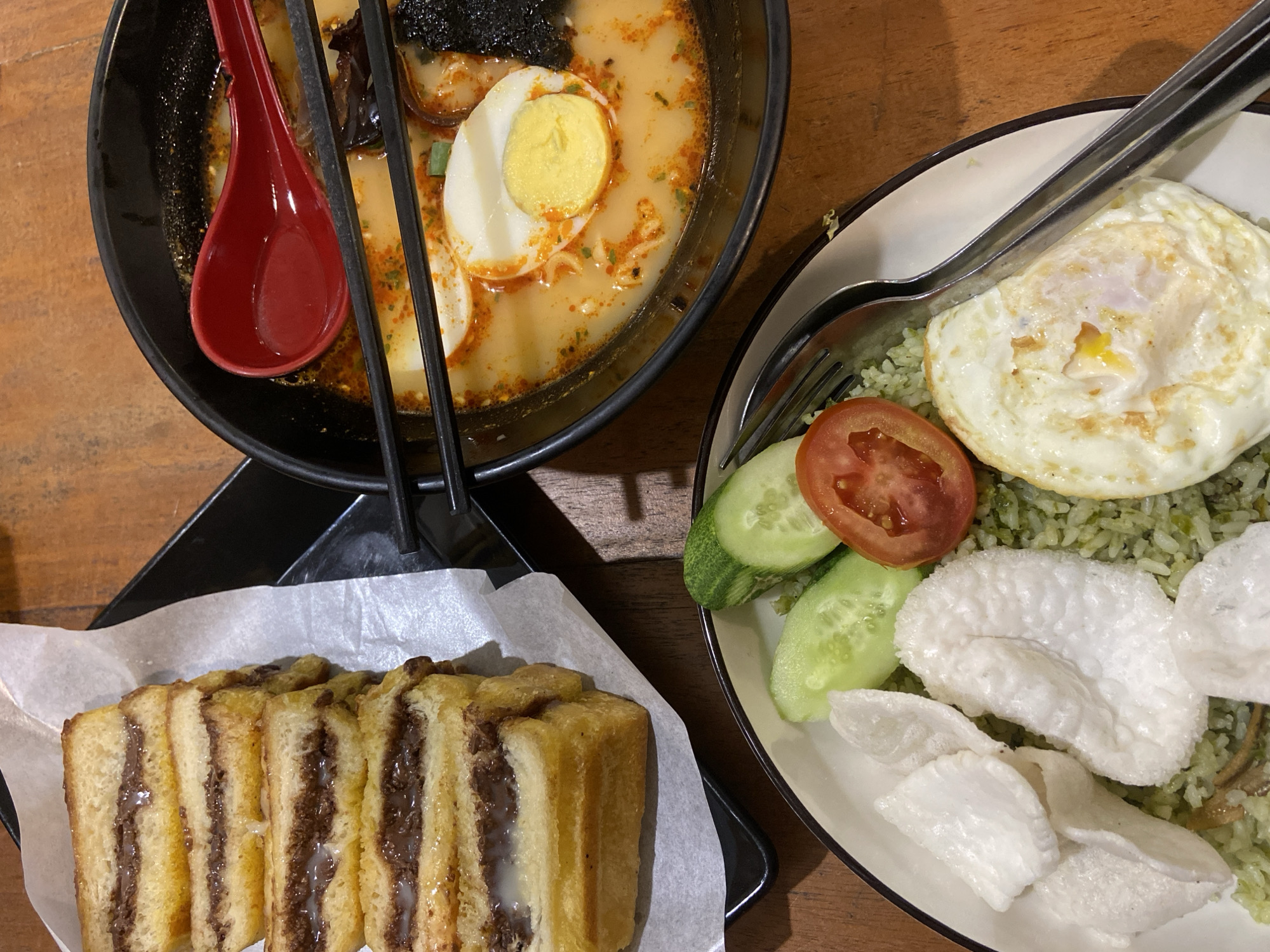 An overhead shot shows a meal arranged on a wooden table. To the left, stacked slices of toasted bread filled with chocolate spread and condensed milk are served on a white paper liner atop a black rectangular plate. In the center, a white plate holds green fried rice topped with white crackers, sliced cucumber, and a slice of tomato, with a fried egg prominently featured on the right side. To the right, a black bowl contains a steaming bowl of ramen with sliced hard-boiled eggs, seaweed, and a red spoon resting inside. Black chopsticks are positioned across the ramen bowl. The scene is indoors, suggesting a dining establishment in Sekar Mawar, Indonesia, given the inclusion of specific Indonesian dishes.