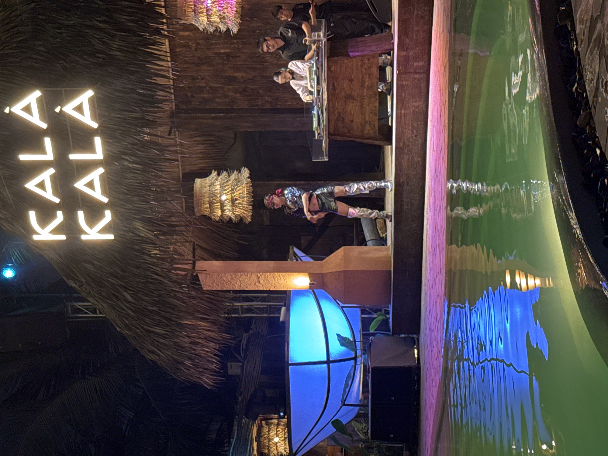 The image shows an outdoor scene at night in Đà Nẵng, Vietnam, likely at a beach club or bar called "Kala Kala". The name is spelled out in bright neon letters on a thatched roof.
A DJ booth is set up with three individuals operating equipment. A female dancer, wearing a sparkly top, dark shorts, and thigh-high boots, is performing below the booth.
A blue light fixture is positioned near what appears to be the edge of a pool or water feature, reflecting blue light. The water is green and reflects other light sources. The overall environment suggests a lively, upscale nightlife setting.