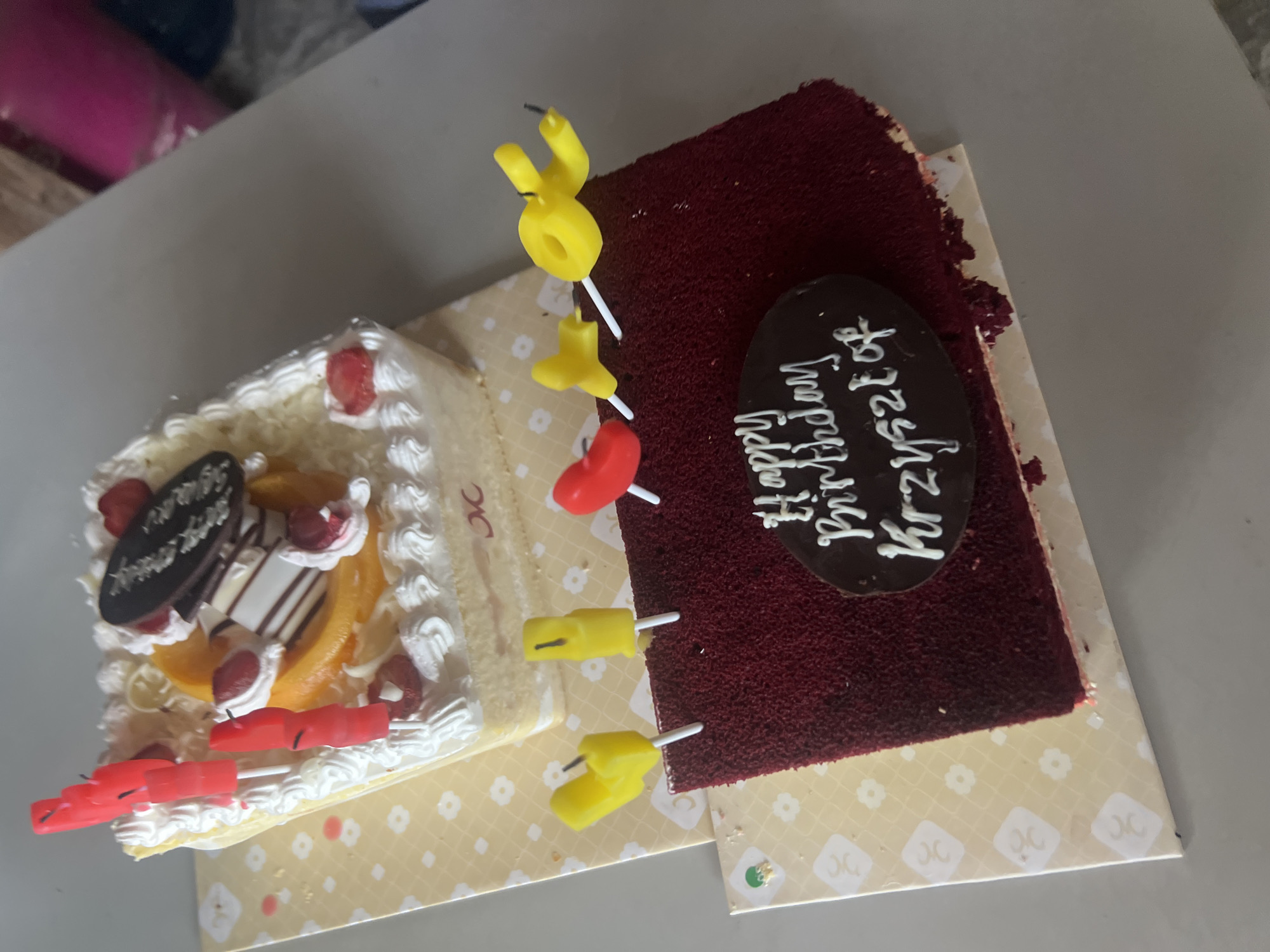 The image shows two cakes on a table. The top cake is a white cake with red candles, fruit slices, and chocolate decorations. The bottom cake is a rectangular red velvet cake with candles and a chocolate oval on top that reads "Happy Birthday Krzysztof". The cakes are on a table with a patterned tablecloth. The setting is not specified in the city of Denpasar, Indonesia.
