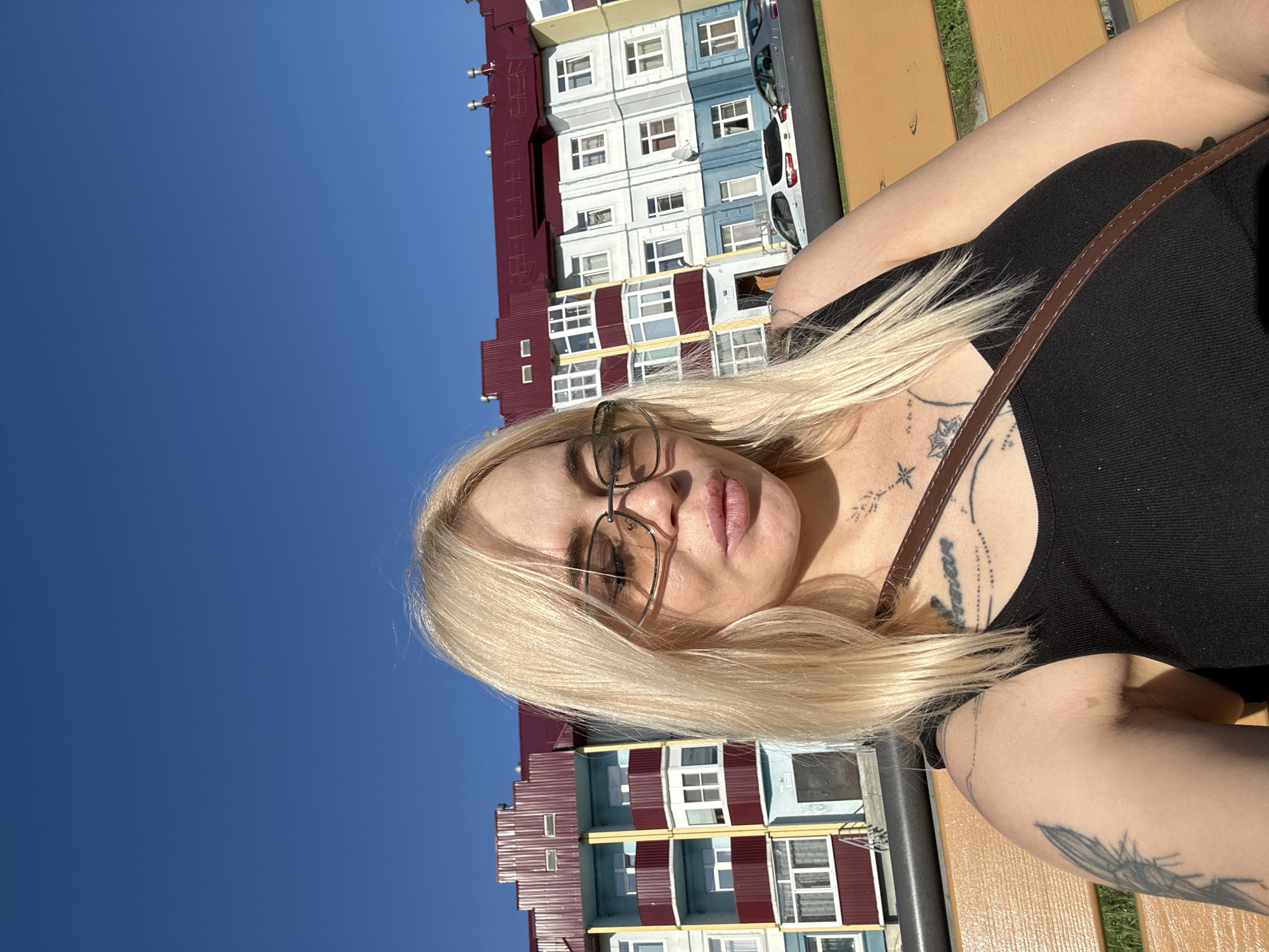 A woman with blonde hair and glasses is sitting on a bench in front of a colorful building. She is wearing a black tank top and has tattoos on her arm and chest. The building is a multi-story apartment building with blue, yellow, and red accents. There are cars parked in front of the building. The sky is clear and blue.