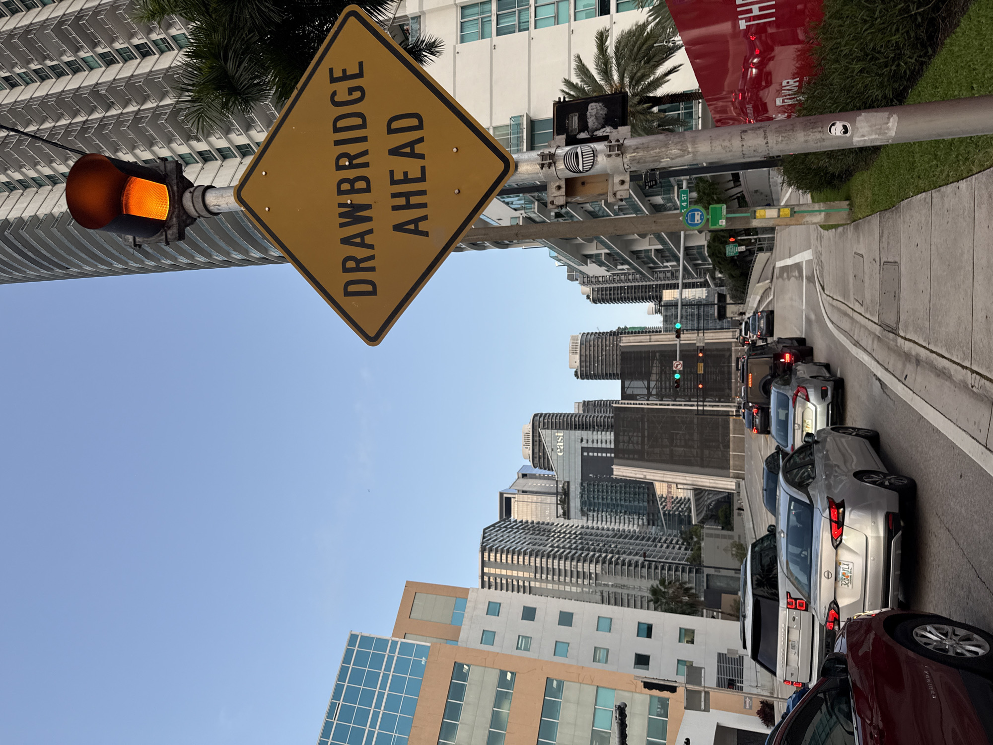 The image shows a city street in Miami, United States of America, with tall buildings and traffic. A yellow "DRAWBRIDGE AHEAD" sign is prominently displayed on a pole next to a traffic light showing yellow. Cars are lined up on the right side of the road, waiting at the traffic signal, with green lights visible further down the road. Buildings of various heights and architectural styles dominate the background. A sticker is seen on the pole holding the traffic light and sign. The sidewalk is visible to the right of the vehicles.