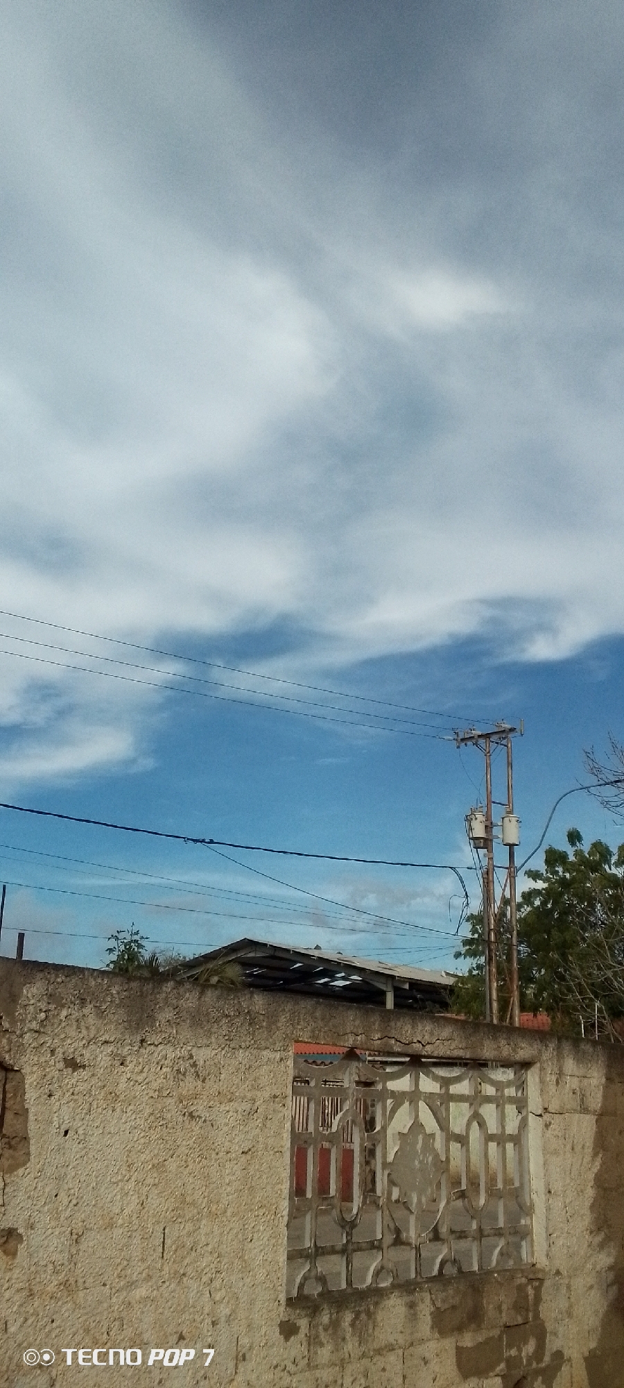 The image depicts a daytime scene in Altagracia, Venezuela. The foreground consists of a weathered concrete wall with a decorative, patterned window or vent. Behind the wall, the corrugated metal roof of a structure is visible. An electrical pole with transformers and power lines crosses the sky. The sky is blue with scattered white clouds. Vegetation can be seen to the right of the electrical pole. The "TECNO POP 7" watermark appears in the lower left corner of the frame.