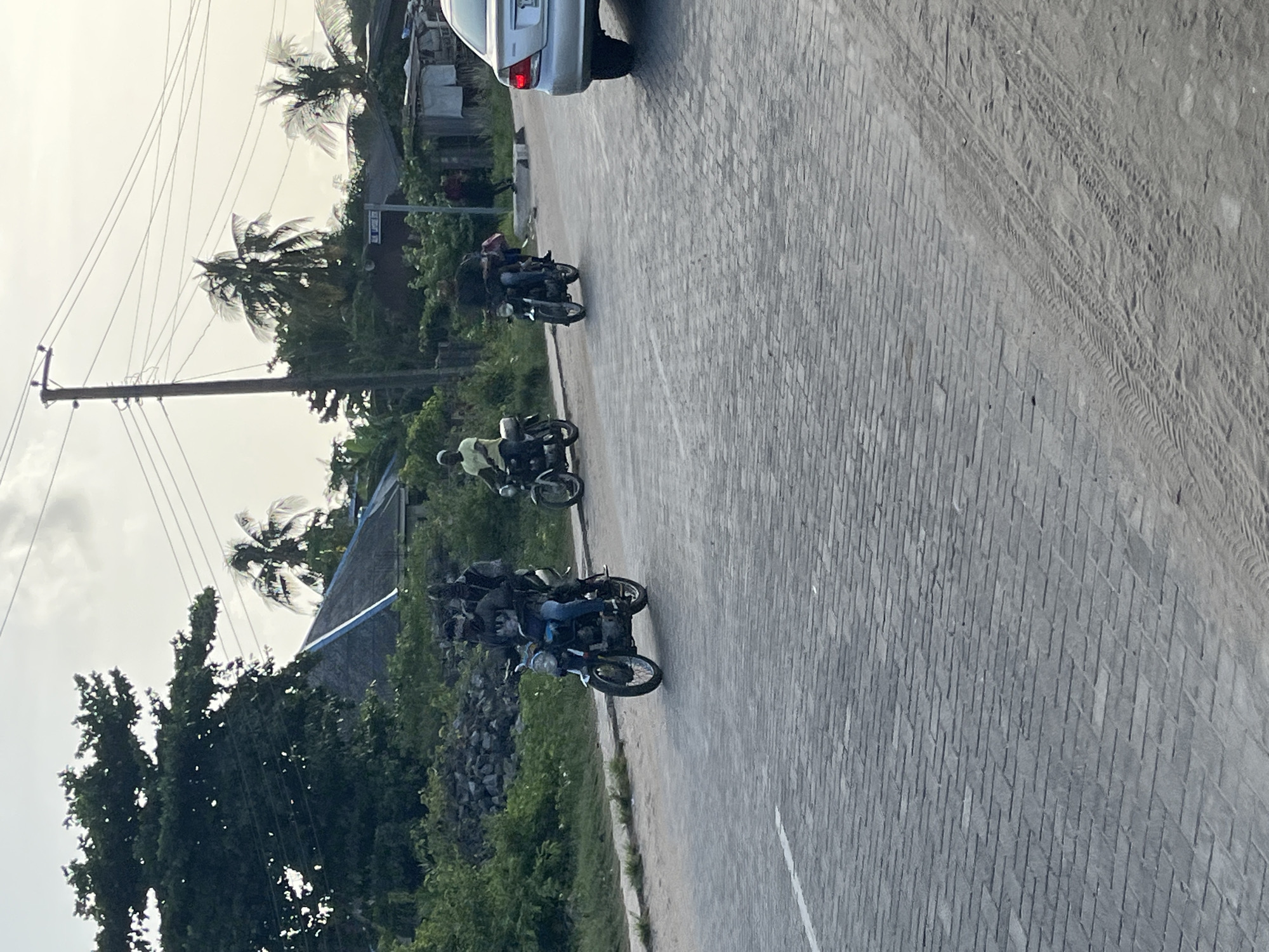 The image depicts a street scene in what appears to be Badagry, Nigeria, given the presence of palm trees and architectural elements. The street is paved with rectangular bricks, and tire tracks are visible on the right side. Three motorcycles are positioned on the left side of the road, each with at least one rider. The riders appear to be local residents. A car is partially visible at the top of the frame. Vegetation lines the side of the road, and a utility pole with wires is visible on the left. The sky is overcast.