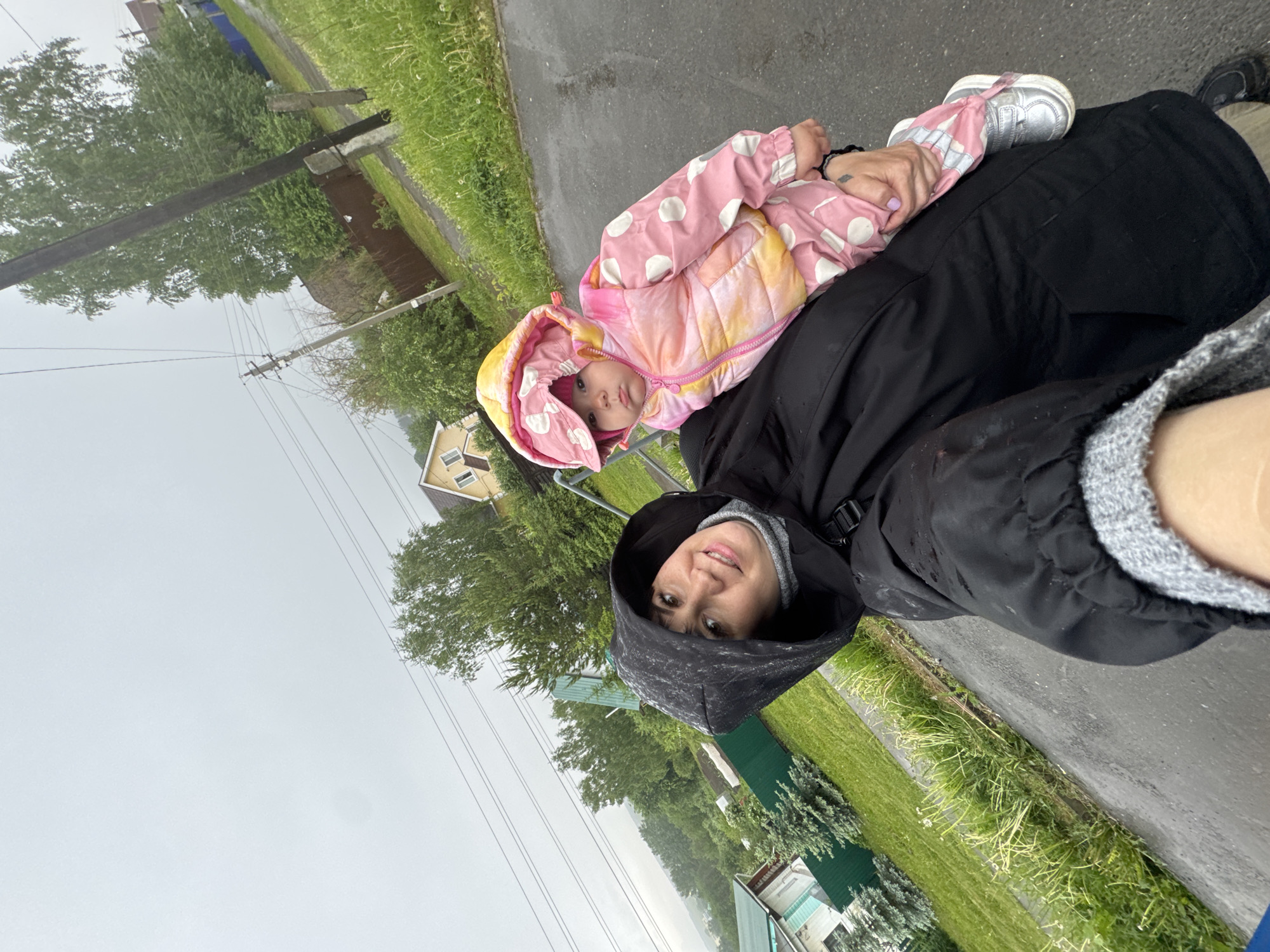 A medium shot shows a woman holding a young child in what appears to be Zav'yalovo, Russia. The woman, wearing a black hooded jacket, smiles into the camera. The child, who is wearing a pink and white polka dot hooded jacket, is reclined against the woman. They are standing on a wet asphalt path with grass and some small buildings visible in the background. Overhead electrical wires are visible. The sky is overcast, suggesting it's either raining or about to rain.