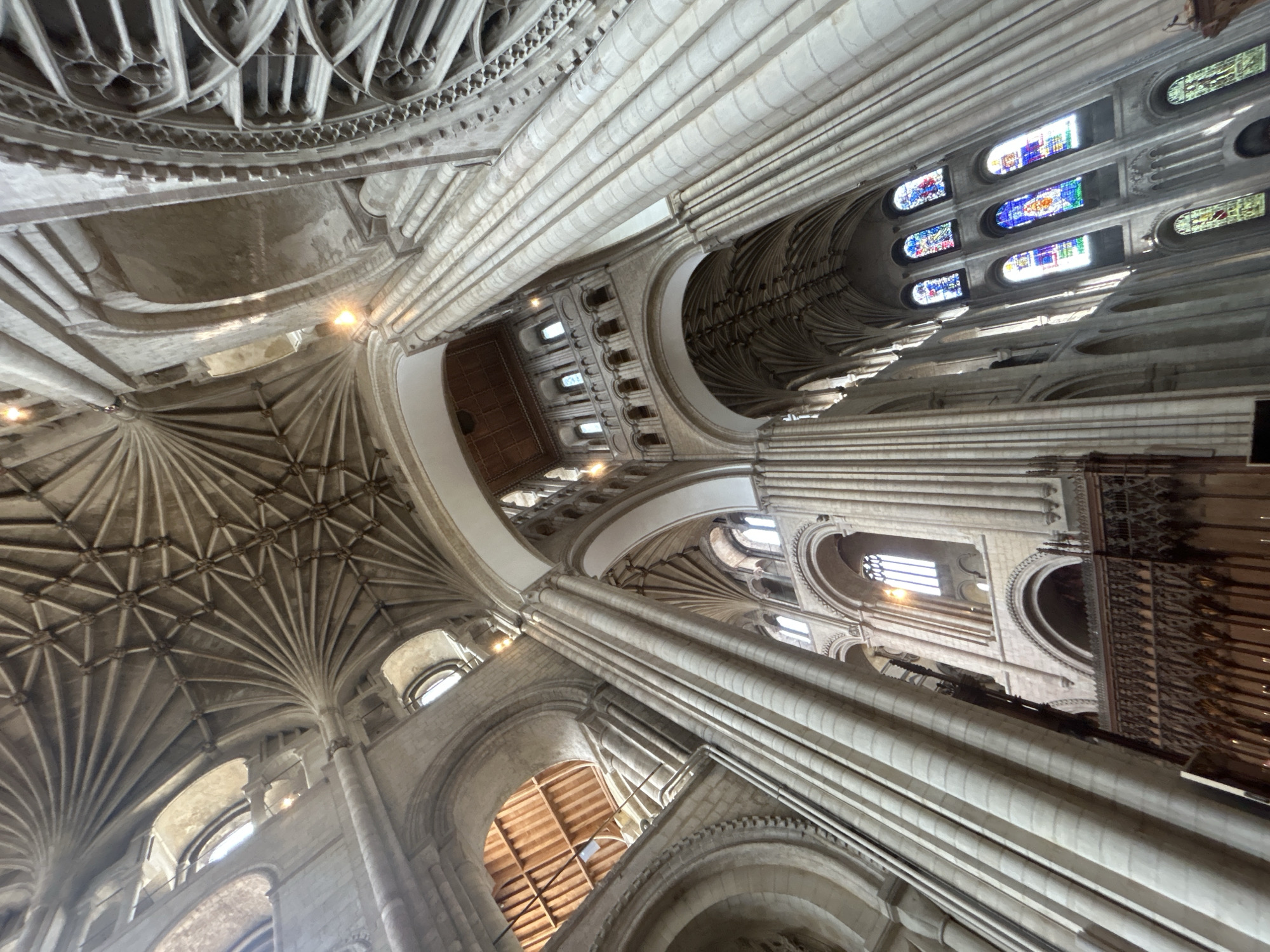 The image shows the interior of Norwich Cathedral in Norwich, United Kingdom. The perspective is from the nave, looking upwards towards the fan vaulted ceiling and the ribbed arches of the triforium and clerestory. Sunlight streams through a large stained-glass window on the right, illuminating colorful panels. Massive stone columns with attached shafts support the soaring architecture. In the foreground, a portion of a carved wooden screen is visible, possibly part of the rood screen. The overall impression is of immense scale, intricate stonework, and the grandeur of Gothic architecture.