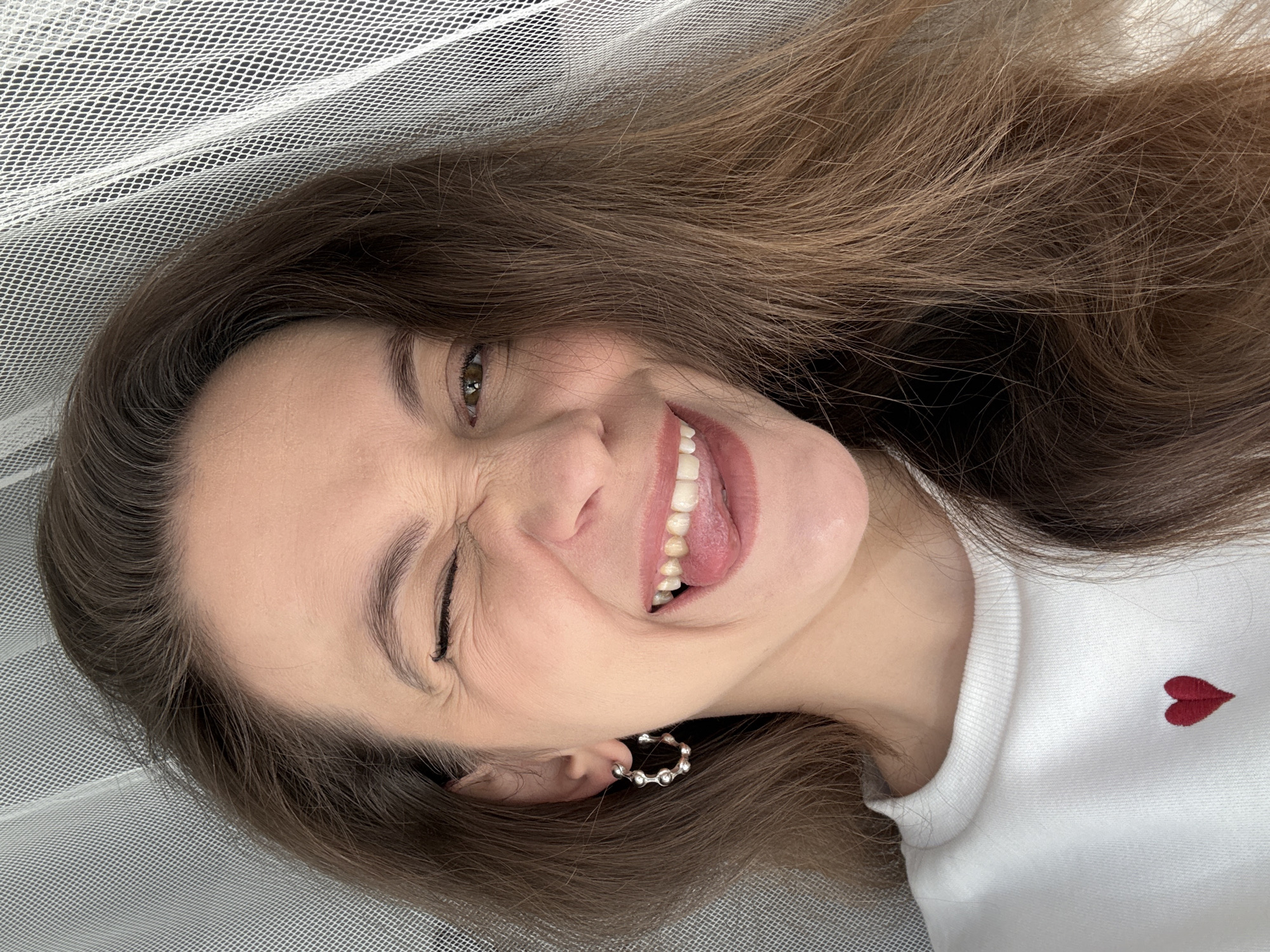 A woman with brown hair and a white shirt with a small red heart on the upper right side of the shirt winks at the camera. She is smiling and her tongue is slightly visible. She is wearing a silver earring in her left ear. She is lying on a white net-like fabric. The location is not discernible from the image.
