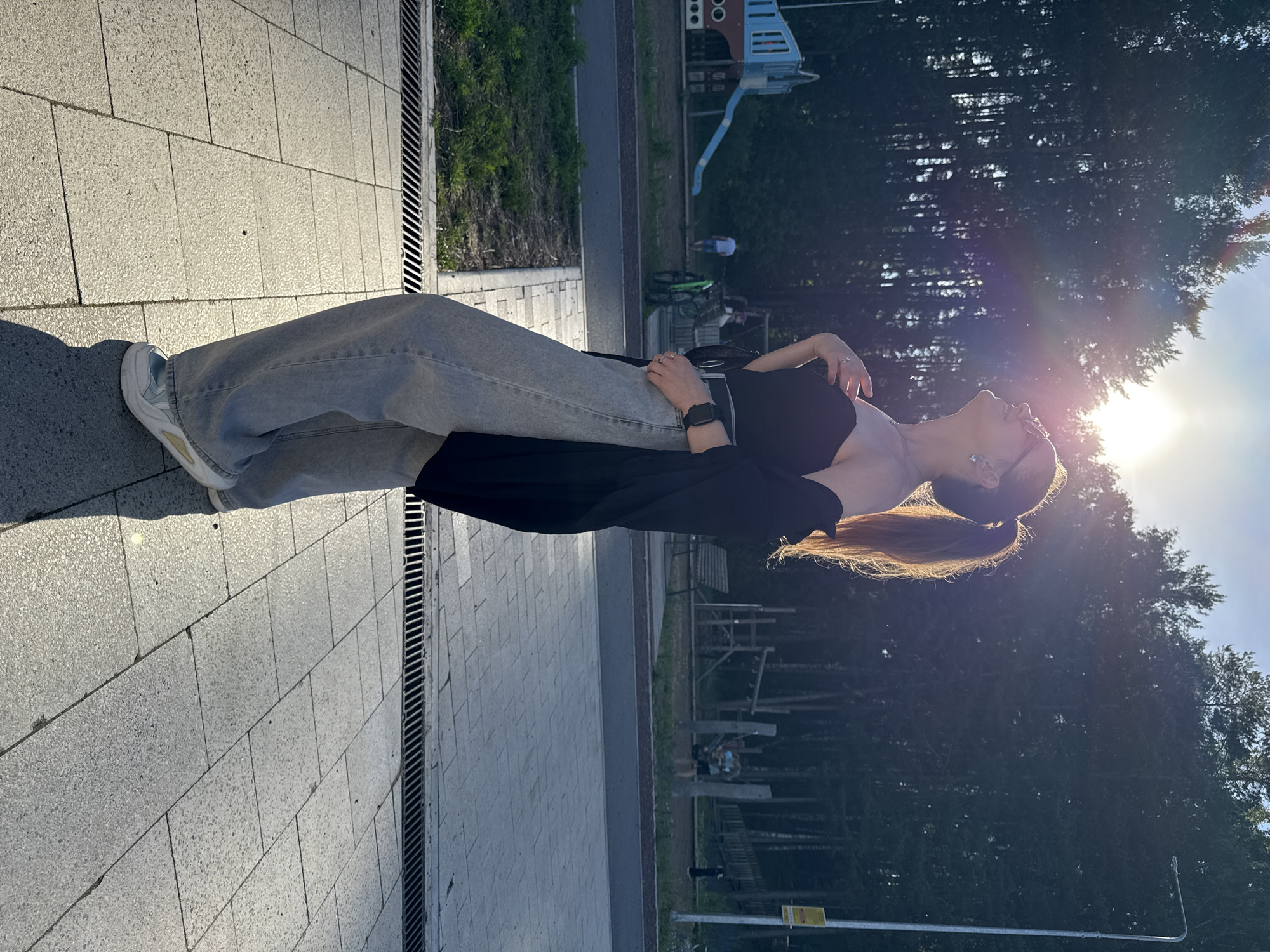 A young woman with her hair in a ponytail stands outside, looking up at the sun. She is wearing a black off-the-shoulder top, light-wash wide-leg jeans, and white sneakers. A black cardigan is draped over her shoulders. She has rings on her fingers and is wearing glasses. The background shows a playground, trees, and a street lamp. The scene appears to be set in a park or public area in Chaikovsky, Russia, as indicated by the presence of a playground and the overall environment. The sun is shining brightly, creating lens flares and casting shadows on the tiled ground.