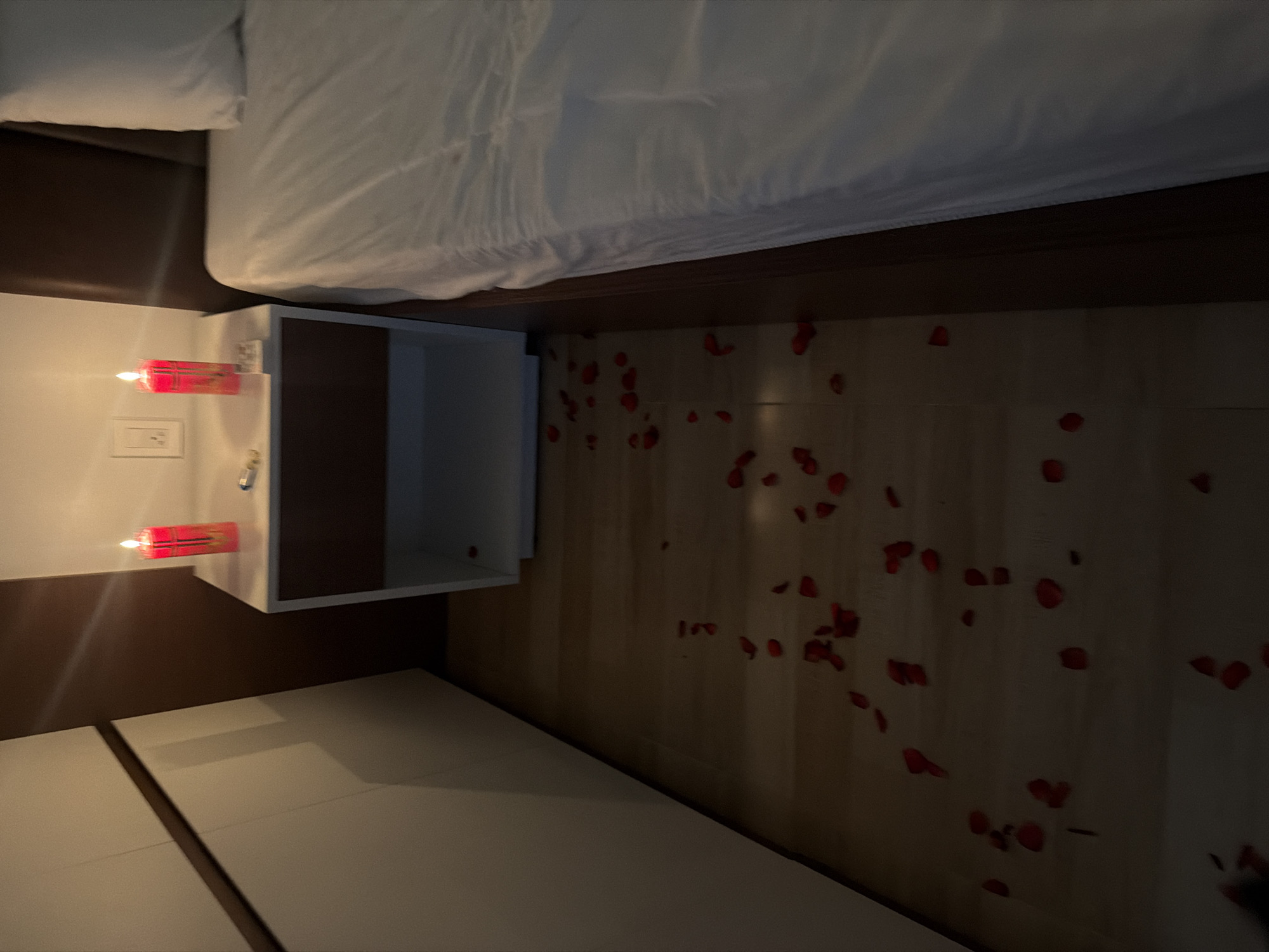 The image depicts a bedroom scene with a romantic ambiance, featuring red candles and rose petals scattered on a wooden floor. In the background, a bedside table holds two lit red candles, one on each side of a light switch. A white bedside cabinet is positioned next to it, and a large bed with white linens is visible to the right. To the left of the frame, a portion of a white wardrobe with dark trim is present. The overall impression is that of a prepared romantic setting, possibly for a special occasion. The image does not provide specific information to indicate it is located in Cam Lam, Vietnam.
