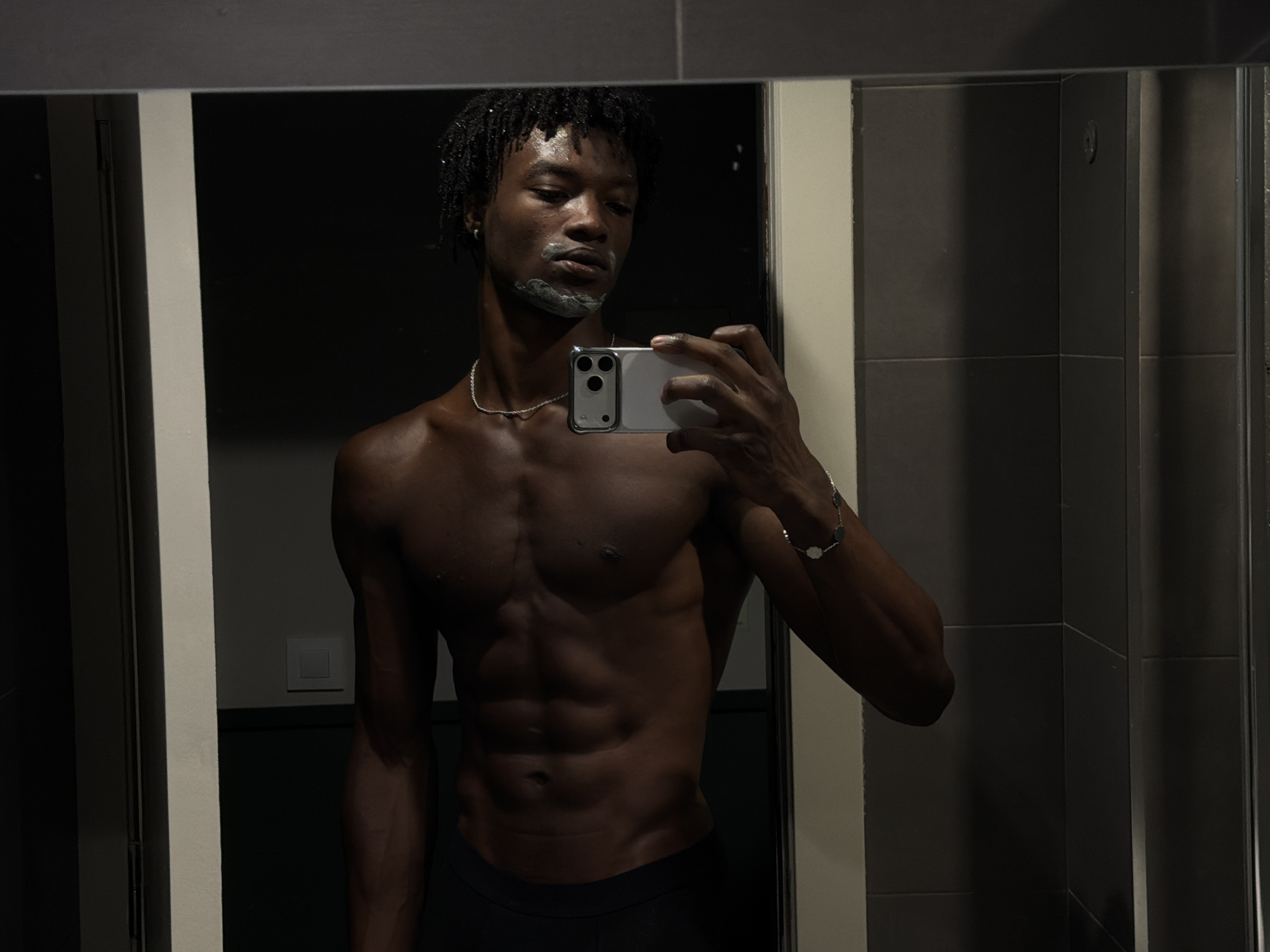 In a dimly lit bathroom, a young Black man with dreadlocks stands before a mirror, taking a selfie with his phone. He is shirtless, displaying well-defined abdominal muscles, and has a thin layer of white shaving cream on his chin and upper lip. He is wearing a silver chain around his neck and a bracelet on his left wrist. The mirror reflects the man, his phone, and parts of the bathroom environment, which includes tiled walls and what appears to be a light switch on the wall. The lighting in the scene is low, with shadows accentuating the contours of his body and the textures of the bathroom. There is no indication of the image being taken in Barcelona, Spain.