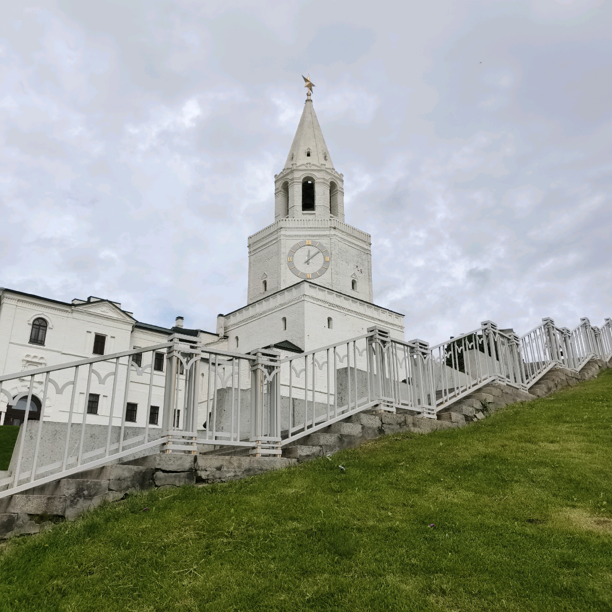 This image depicts the Söyembikä Tower within the Kazan Kremlin, a UNESCO World Heritage Site located in Kazan, Russia. The tower, a prominent landmark, is constructed of white brick and features a distinctive clock face with Roman numerals. A golden star adorns its spire. In the foreground, a wide, white-railed staircase ascends a grassy hill, leading towards the Kazan Kremlin complex. The sky above is overcast with clouds, and the overall scene is a daytime shot with a slightly muted color palette. There are no people visible in the frame.