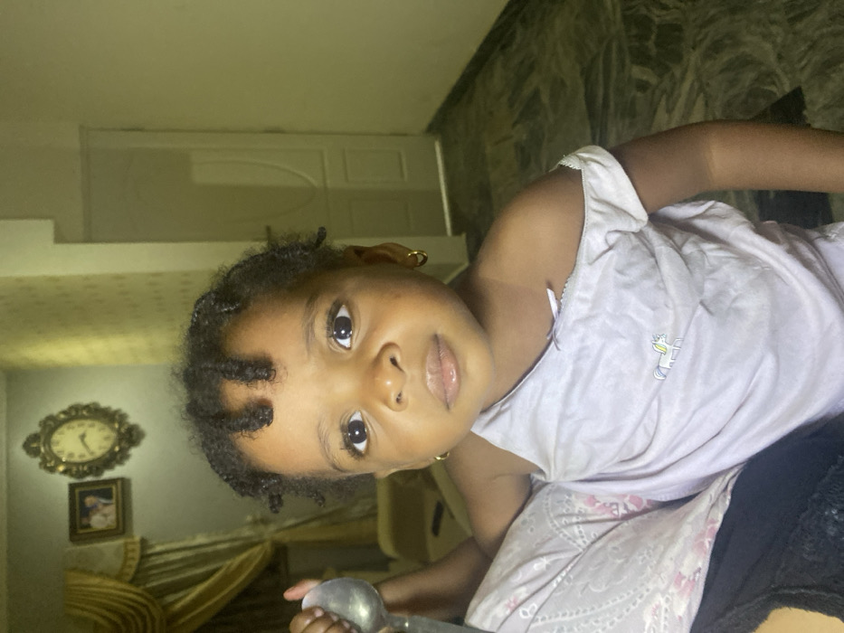 A young child with dark, curly hair and gold hoop earrings looks directly at the viewer. The child is wearing a light purple shirt adorned with a small unicorn. The child is holding a silver spoon. In the background, there is a white door, a clock, framed picture, and a curtain. The floor is marble.