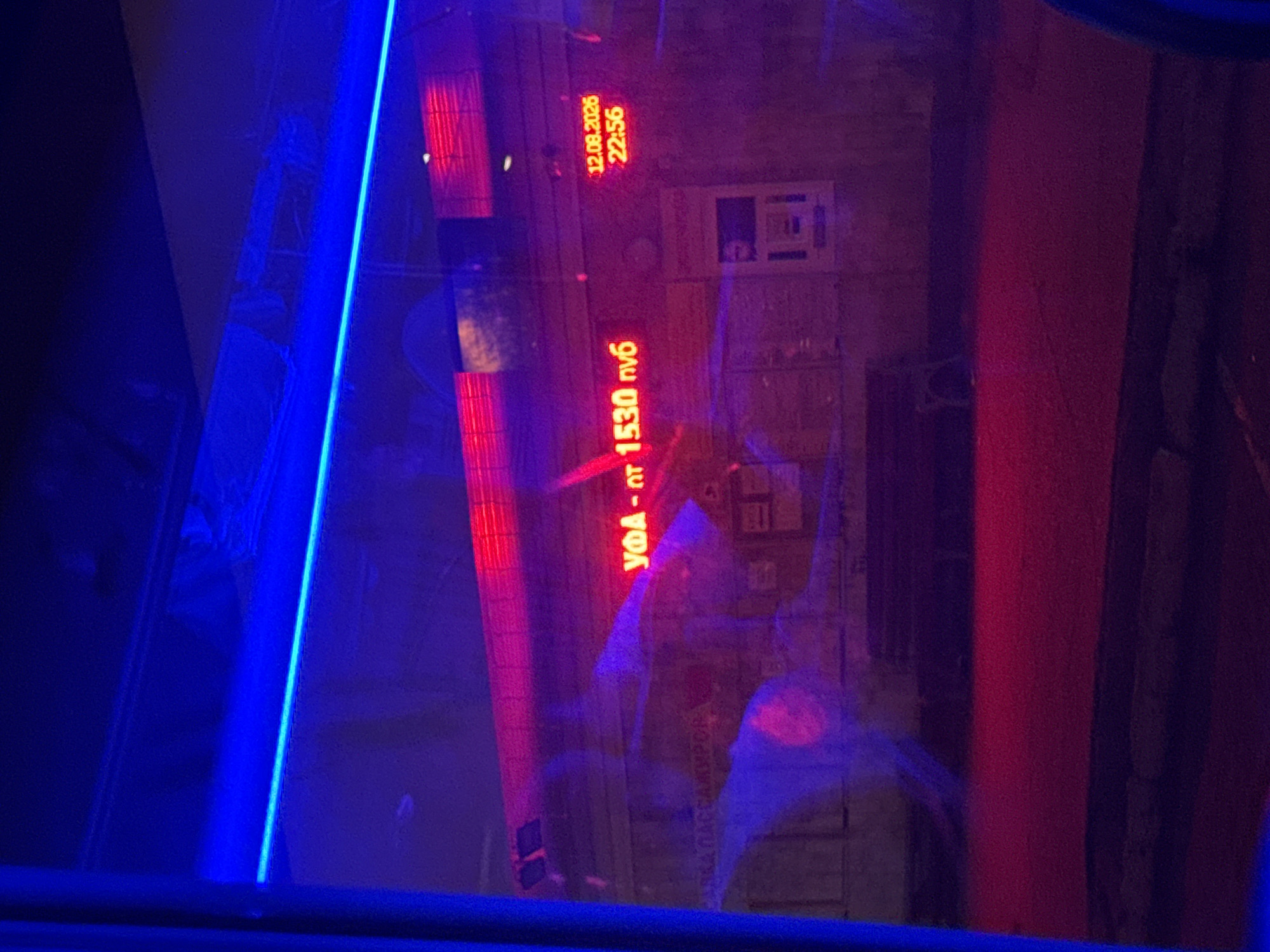 The image shows a night scene in Podgornyi Driush, Russia, viewed through a reflective surface, likely a window. Neon lights dominate the scene, with a prominent blue light strip running horizontally across the top, and red neon lights illuminating a building facade. A red digital display on the building shows "УФА - от 1530 руб" (UFA - from 1530 rubles) and another display shows "12.08.2026 22:56". The building appears to be a bus station or similar public transport facility, as indicated by a partially visible sign that reads "ЯРКАЯ ПАССАЖИРОВ" (likely part of "ЯРКАЯ ПАССАЖИРСКАЯ" meaning "Bright Passenger"). There is a bench visible in front of the building. The overall impression is one of a somewhat desolate urban environment at night, illuminated by artificial light.