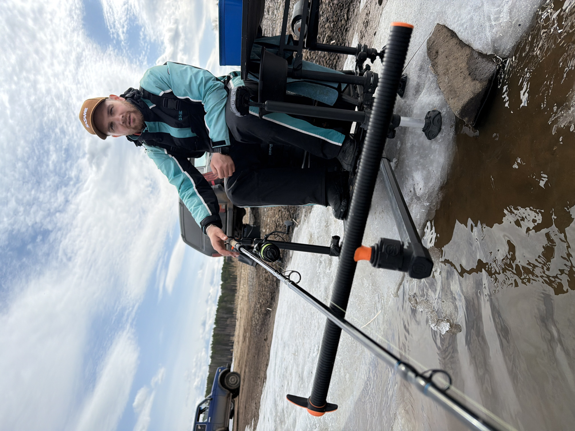 A man in a brown cap and blue and black jacket is fishing on a partially frozen body of water. He is sitting on a black fishing platform. A fishing rod is held in his hand, extending into the water. Patches of ice are visible on the water's surface and along the shore. A rock is visible at the water's edge, partly covered in ice. A blue car is parked in the background on the left side of the frame. The sky is overcast with light clouds. The location is Chaikovsky, Russia.