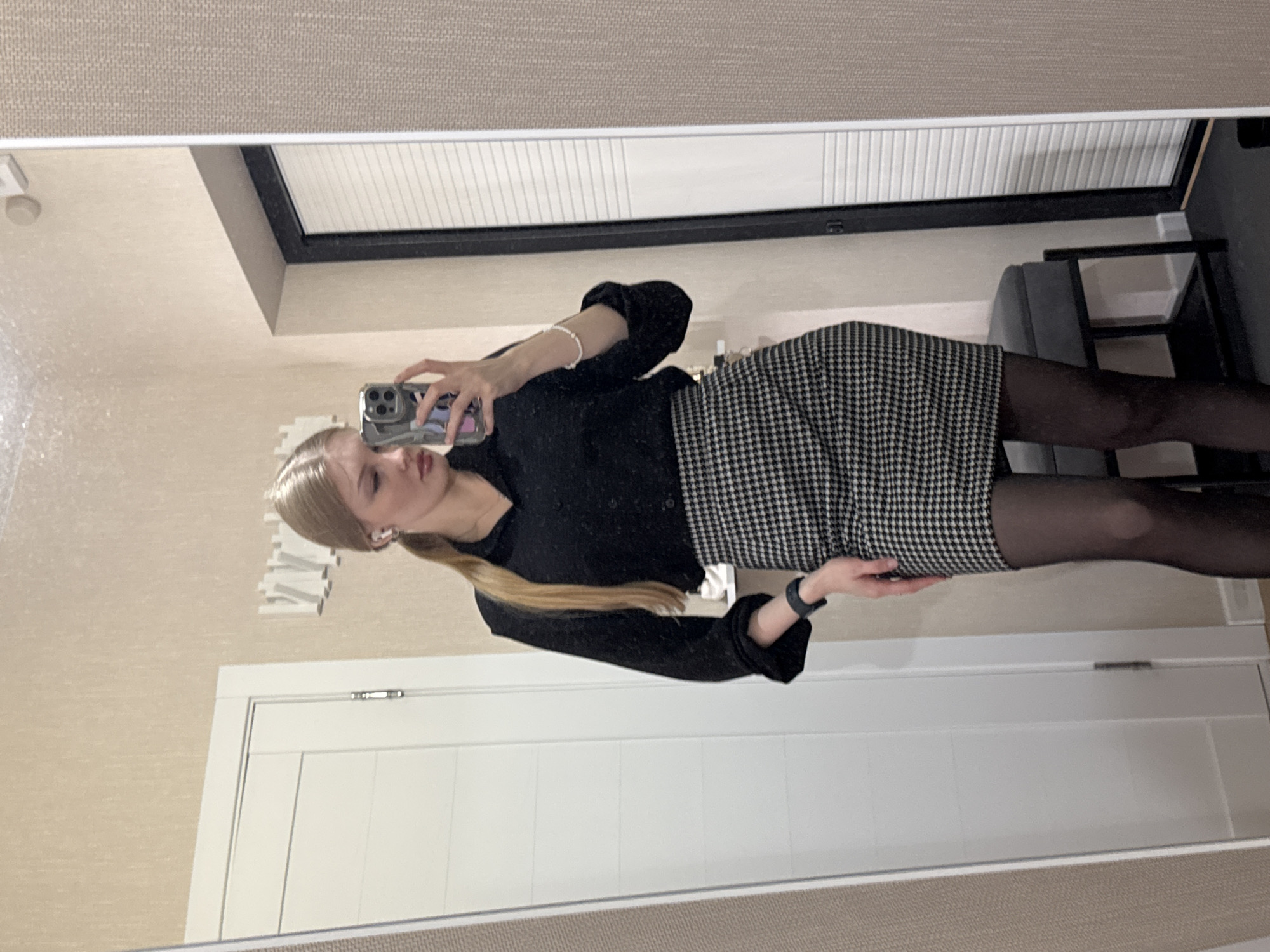 The image shows a young woman with blonde hair taking a selfie in front of a mirror. She is wearing a black blouse, a houndstooth skirt, and black tights. The background includes a white door, a window with closed blinds, a chair, and a beige wall. Based on the analysis of available information, there is no indication this image was taken in the city of Perm, Russia.