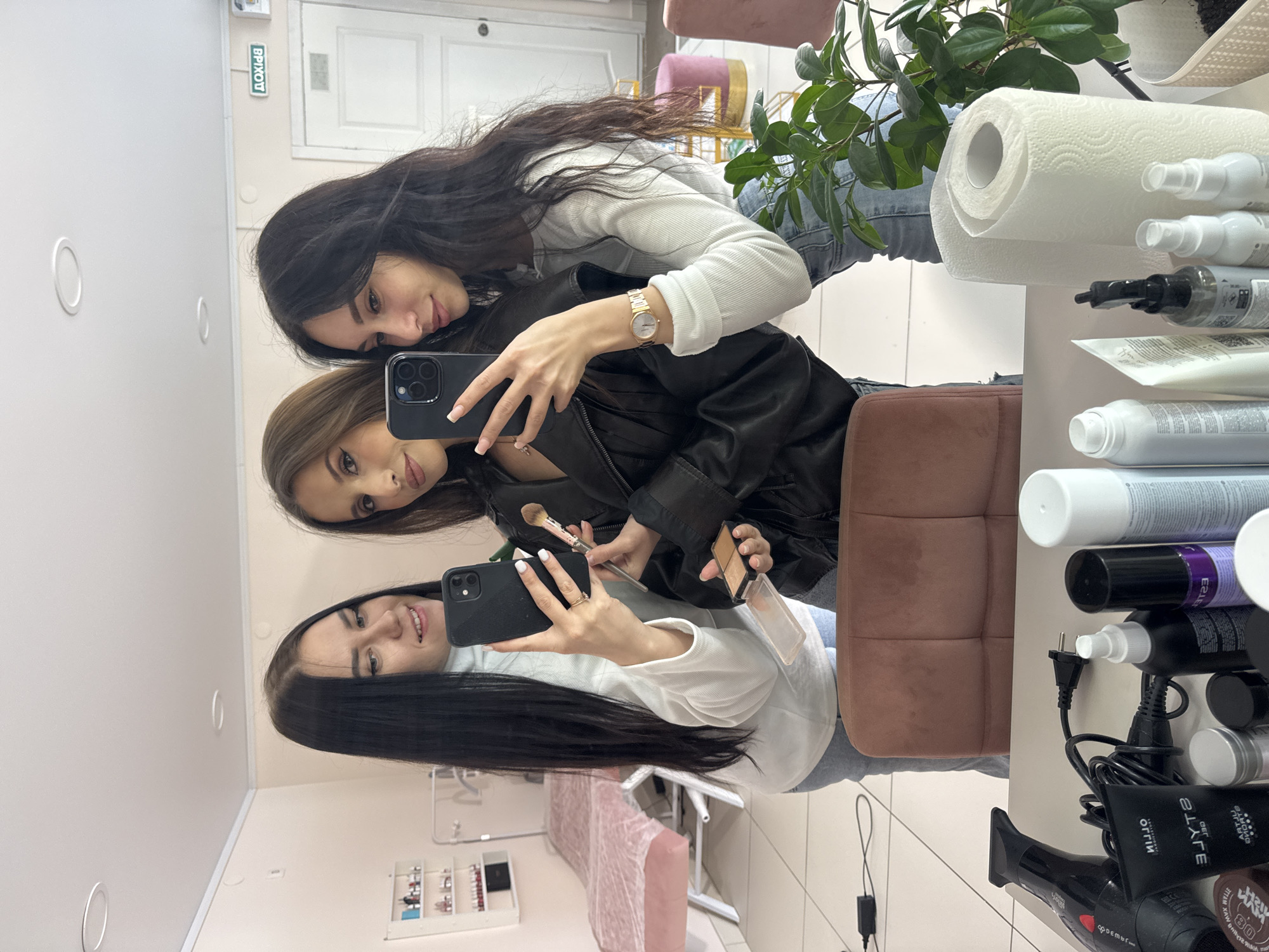 Three women take a selfie in a beauty salon in Chaikovsky, Russia, judging by the “выход” (exit) sign in Russian. The top woman has long, dark hair, wearing a white long-sleeve shirt and a gold wristwatch. The middle woman has light brown hair and is wearing a black leather jacket. The bottom woman has long, dark hair and wears a white long-sleeve shirt. She holds a black iPhone with a makeup brush and an opened makeup palette in her hands. The beauty salon contains typical items: a mirror, chairs, a plant, paper towels, hair products, a hair dryer, and a display case with beauty products on the wall.