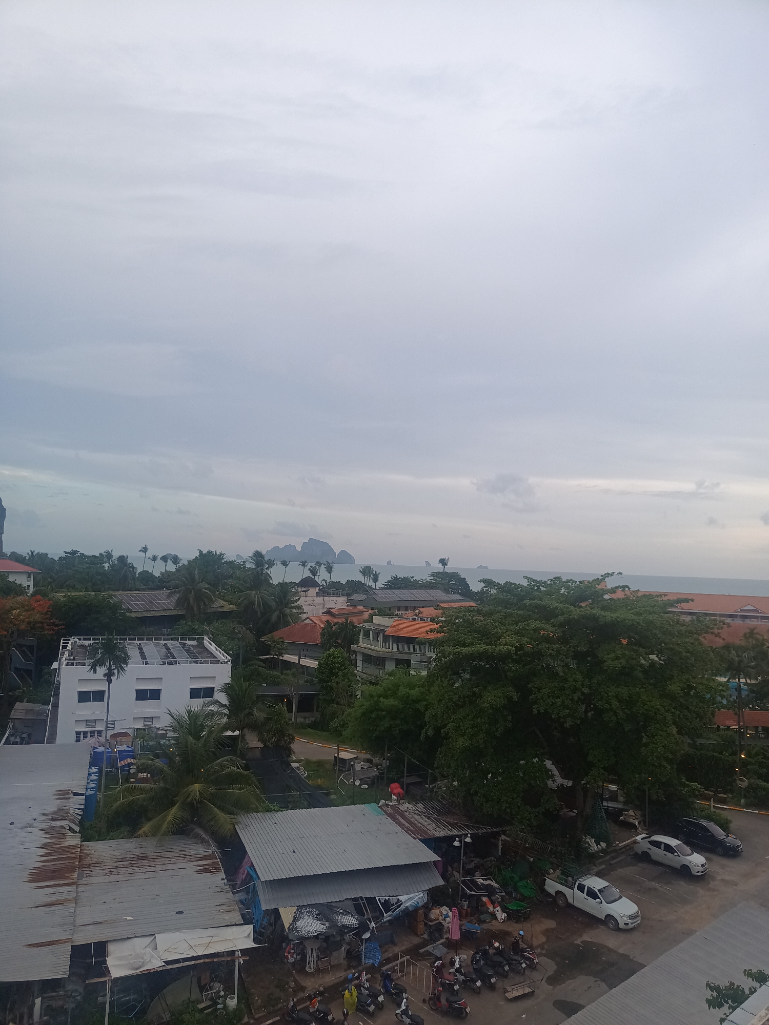 The image shows a view overlooking Ban Ao Nang, Thailand on an overcast day. The scene includes residential and commercial buildings with diverse roof structures, ranging from corrugated metal to red tile. Lush tropical vegetation, including palm trees and large trees, is interspersed among the buildings. In the distance, the coastline is visible with several islands or rock formations rising from the sea. A parking lot is situated in the lower right of the frame, occupied by cars and motorcycles. The sky is predominantly gray, indicating cloudy conditions. The photo appears to have been taken from an elevated position within the city.