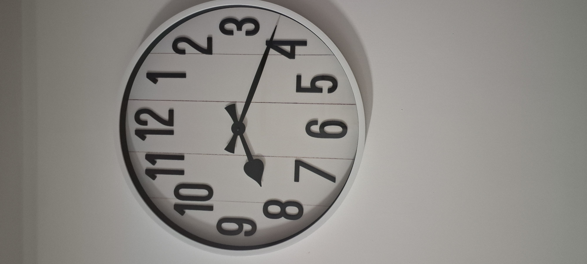 A white circular wall clock with black Arabic numerals hangs on a plain, light-colored wall. The clock has black hour and minute hands. The hour hand points to the number 8, and the minute hand points to the number 4. The wall appears to be indoors, likely in a residential setting. The location is unknown, and it is not possible to determine if this is in Ashkelon, Israel.